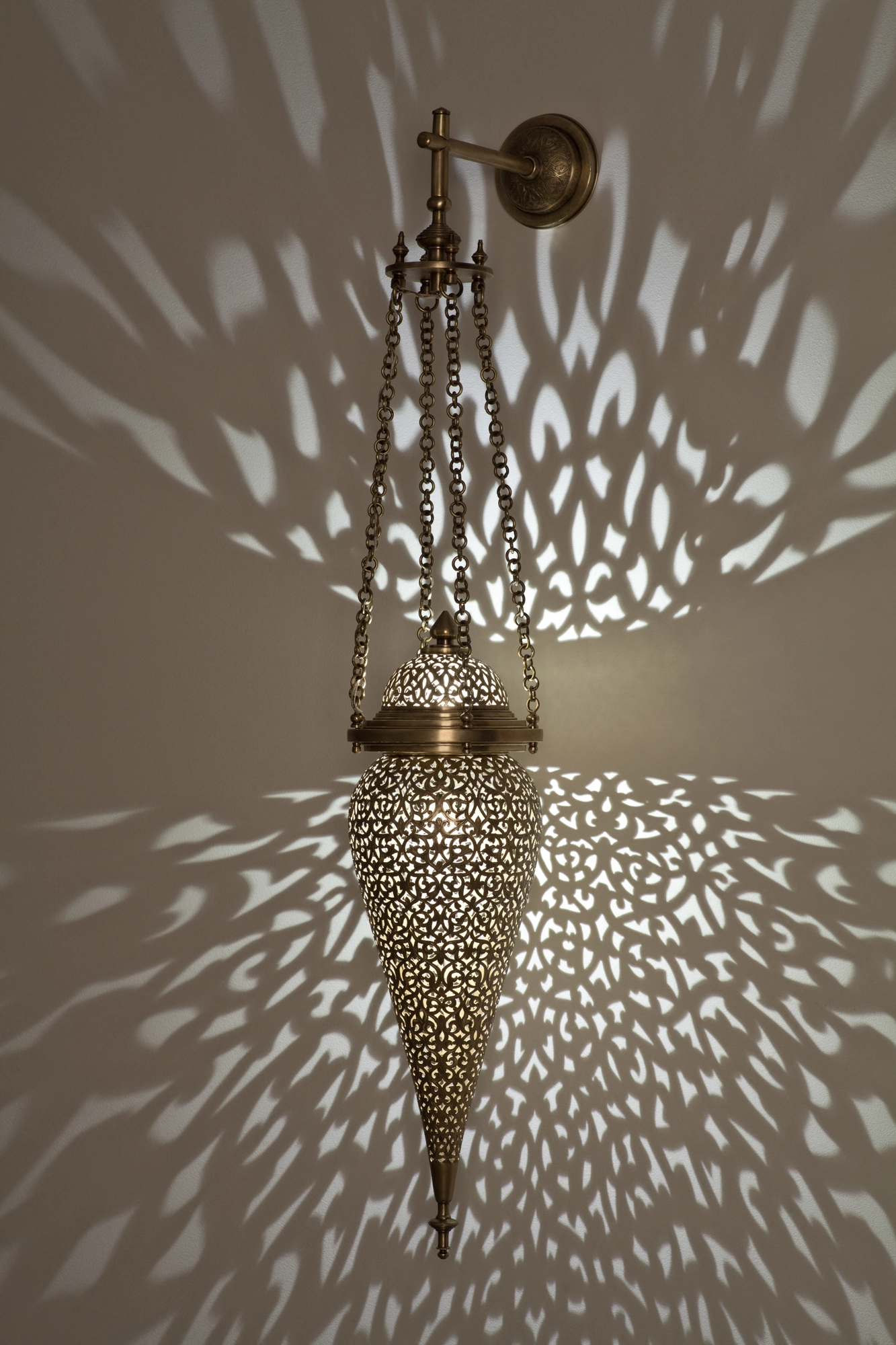 Inverted pierced-brass tear-drop wall light hung from a bracket, glowing within a wide scatter of light dots.