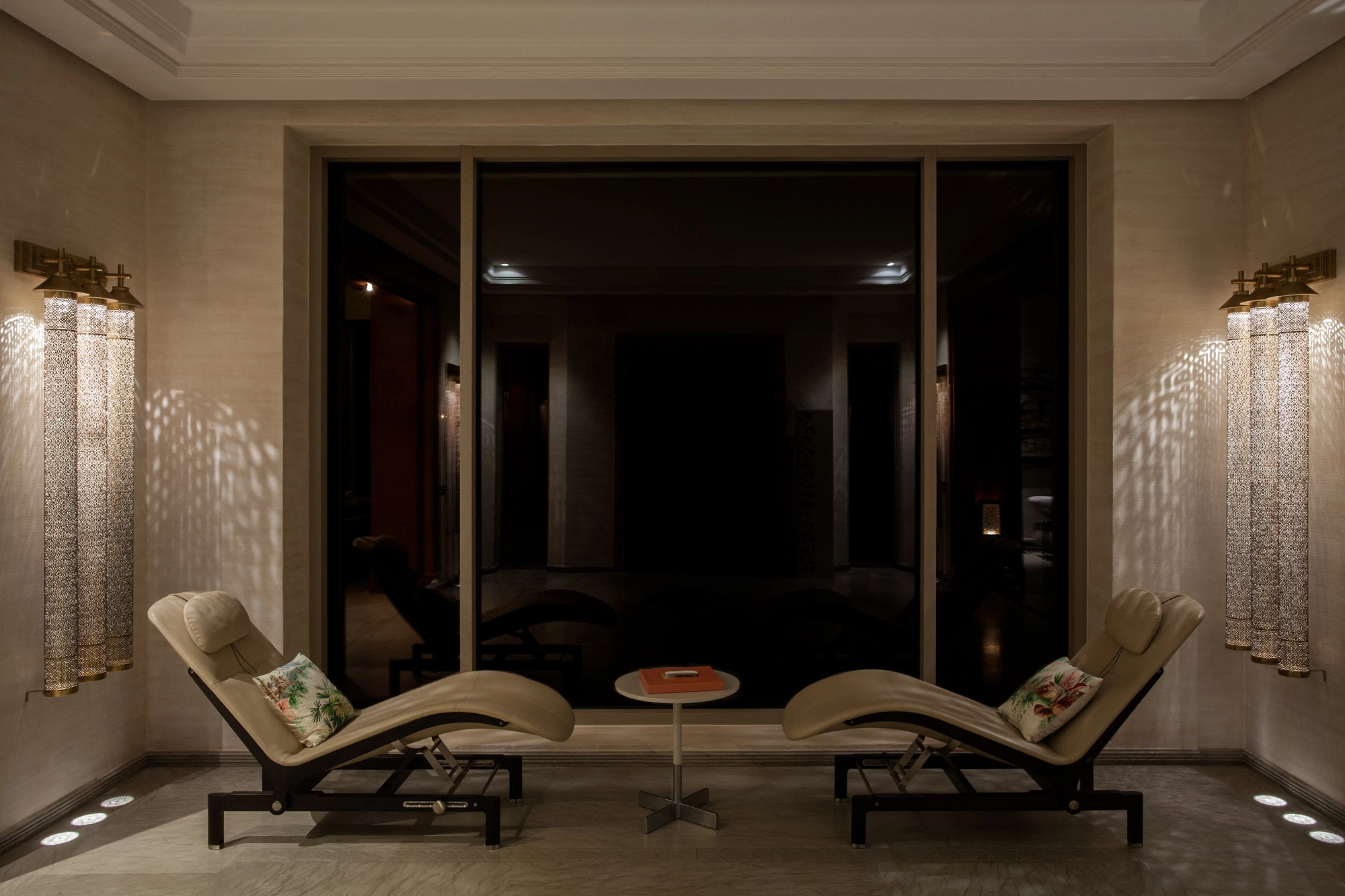 Triple pierced-cylinder wall lights in a dark room with two chaise lounges on a mirror-like floor.