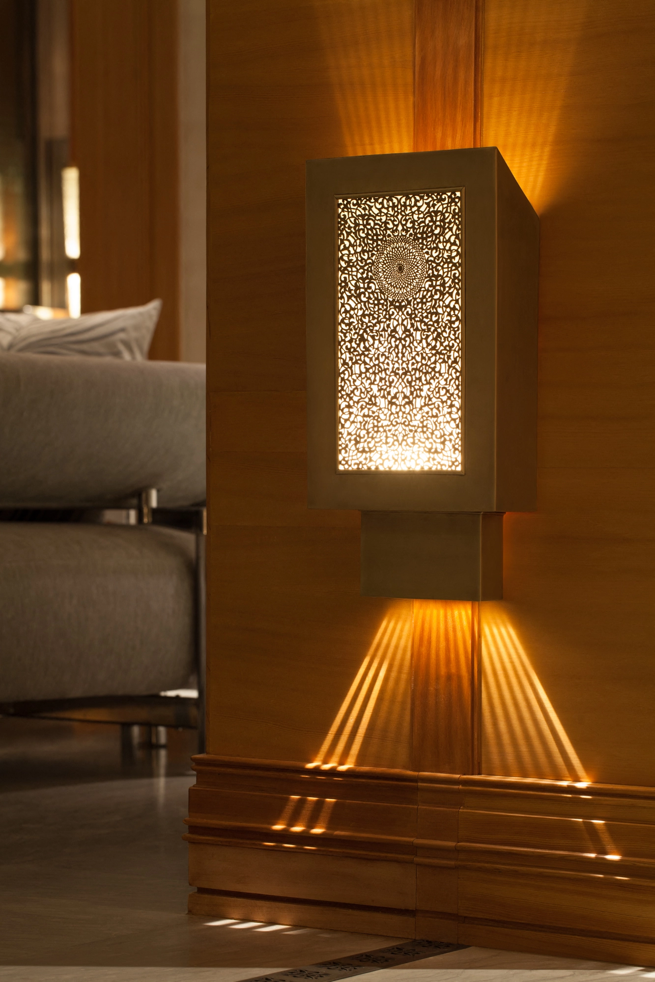 Trapezoidal pierced-box wall light glowing on a wood-panelled wall beside a lounge chair.