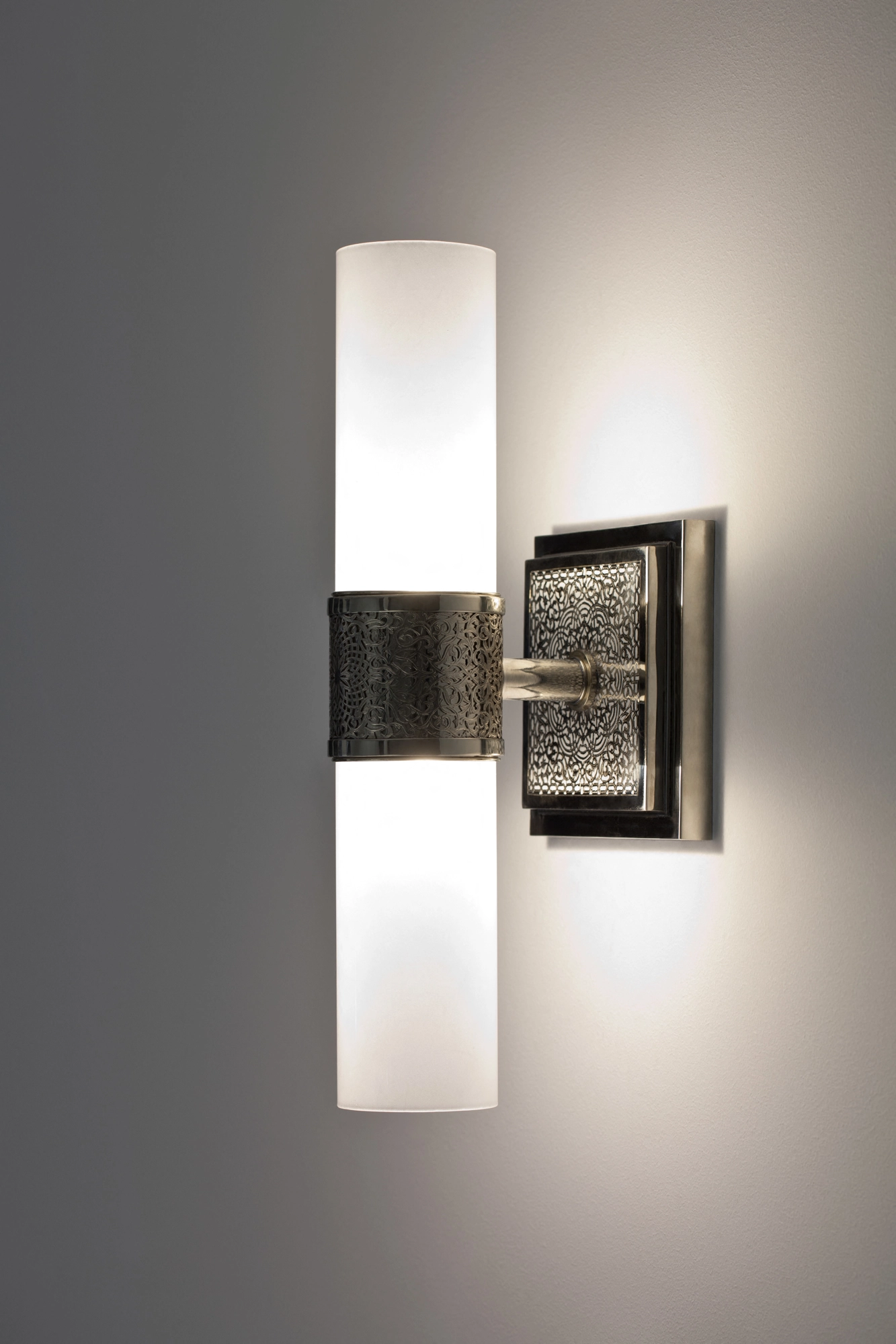 Wall sconce with two frosted glass cylinders on a pierced-metal backplate, glowing softly against a grey wall.