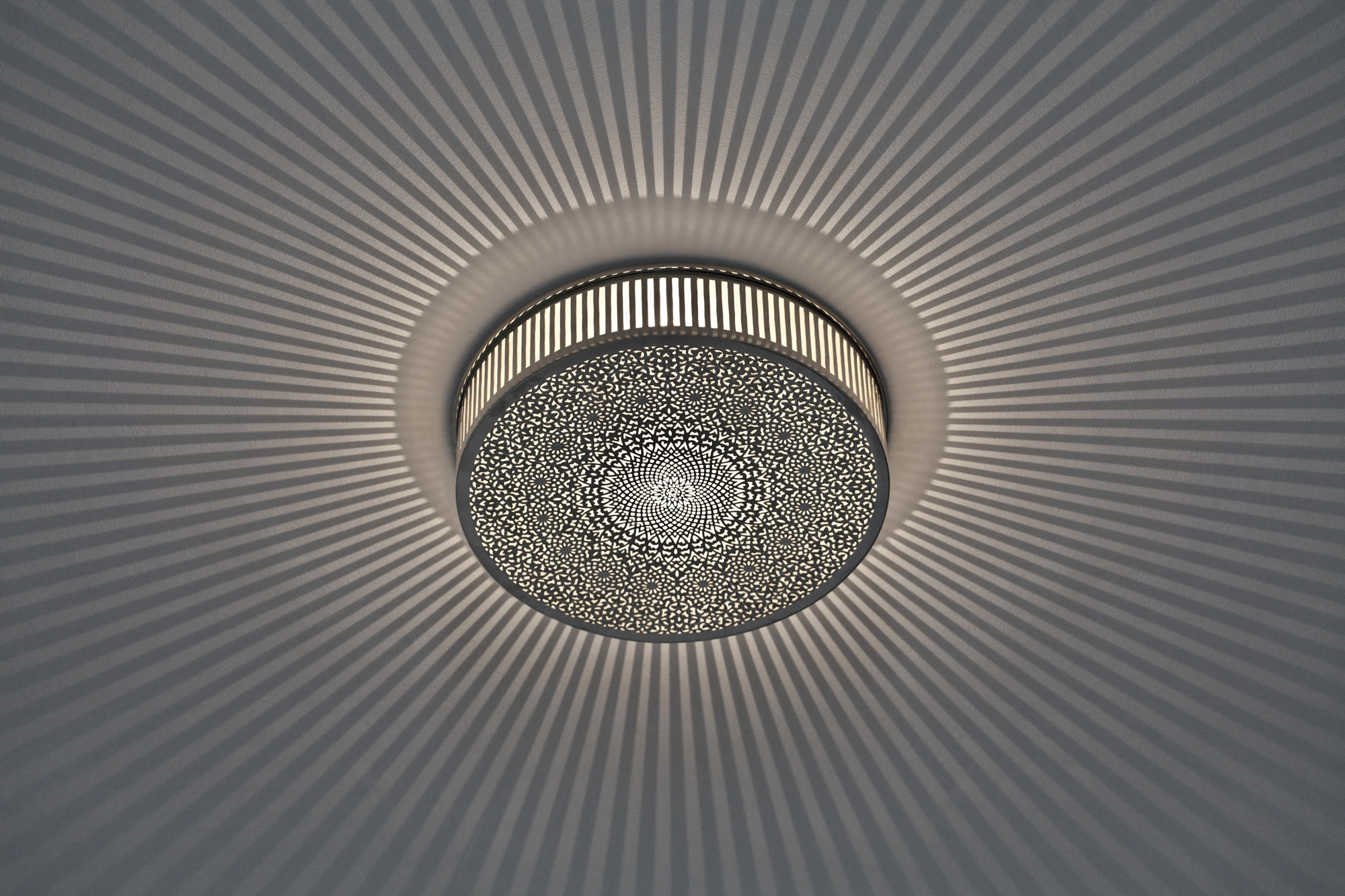 A circular pierced metal rosace ceiling light scattering a fine radial spray of light across a pale grey ceiling.