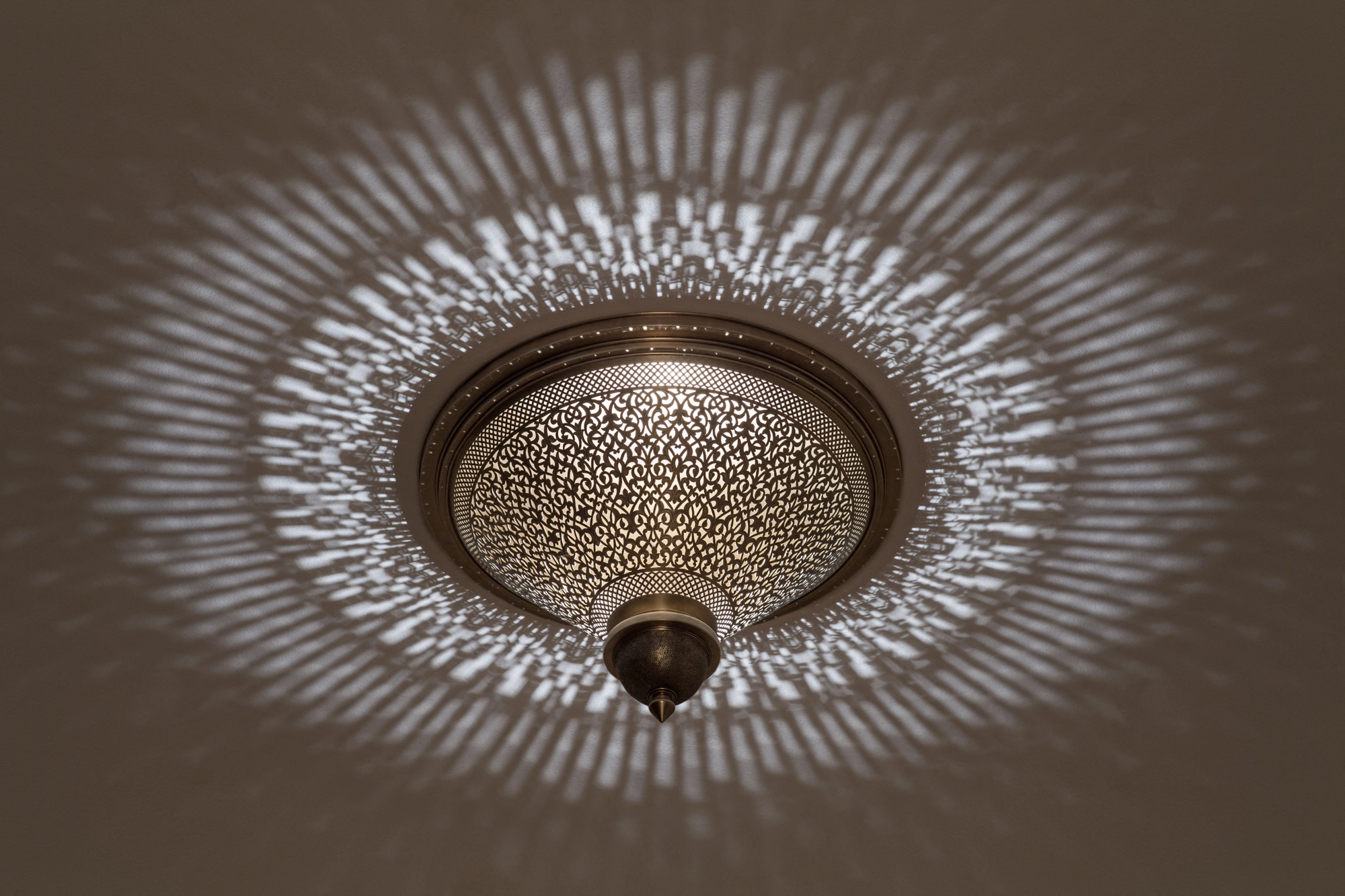 A pierced metal ceiling light seen from below, scattering dense dappled light across a warm-toned ceiling.