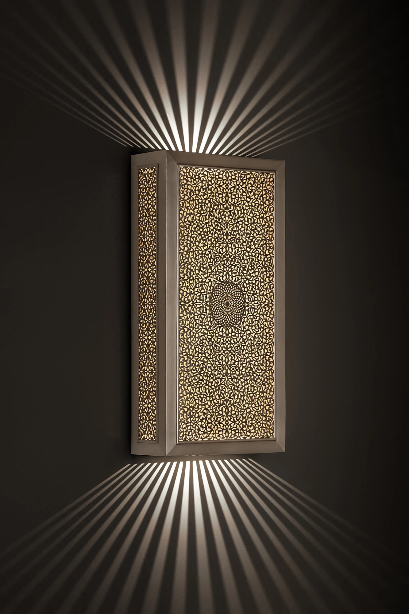 Vertical pierced-brass flat box wall light glowing on a dark wall, casting fans of light upward and downward.