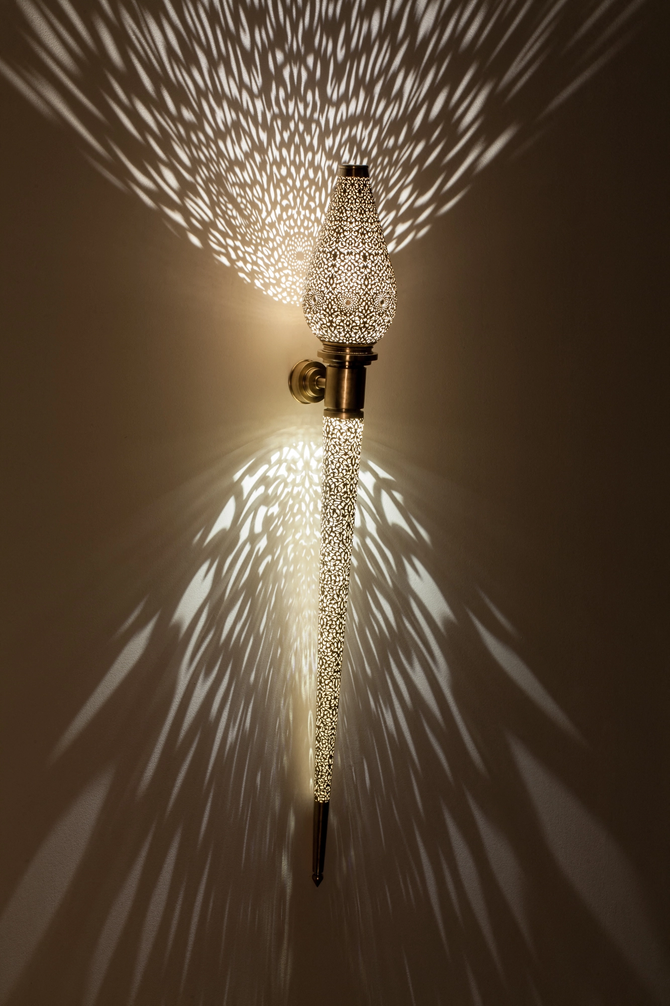 Pierced-brass Treville torch wall light glowing on a bracket, casting feathery rays of light across the wall.