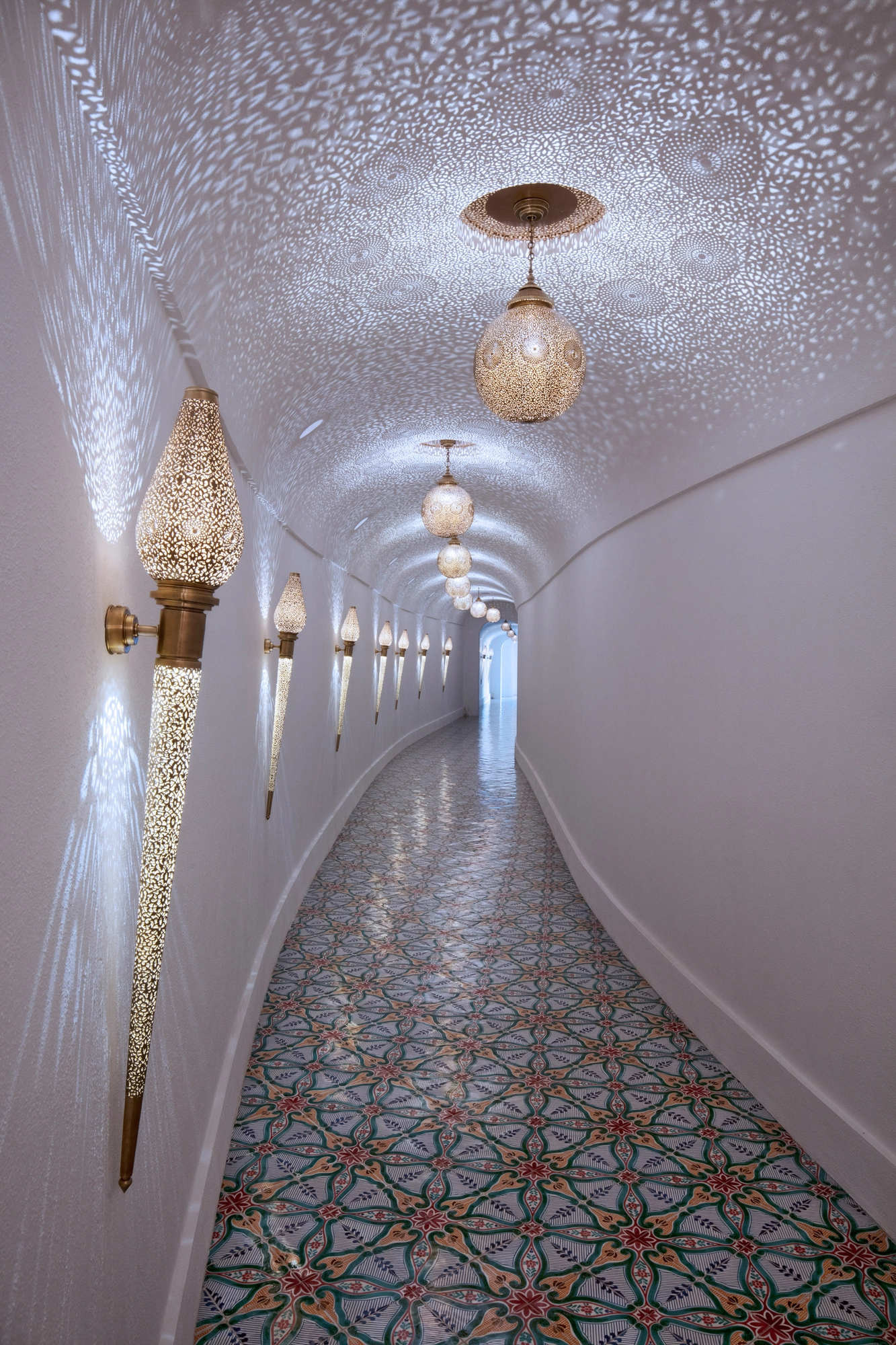 Curved white tunnel corridor with torch wall lights, a hanging lantern and patterned tile floor.