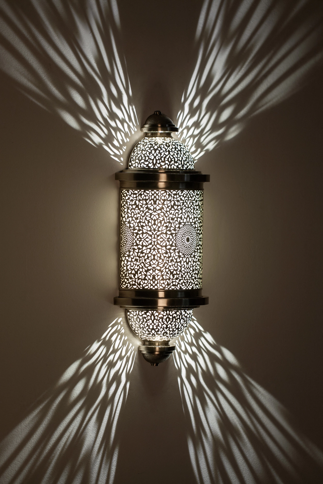 Pierced-brass barrel wall light glowing on a dark wall, throwing an X-shaped burst of light rays outward.