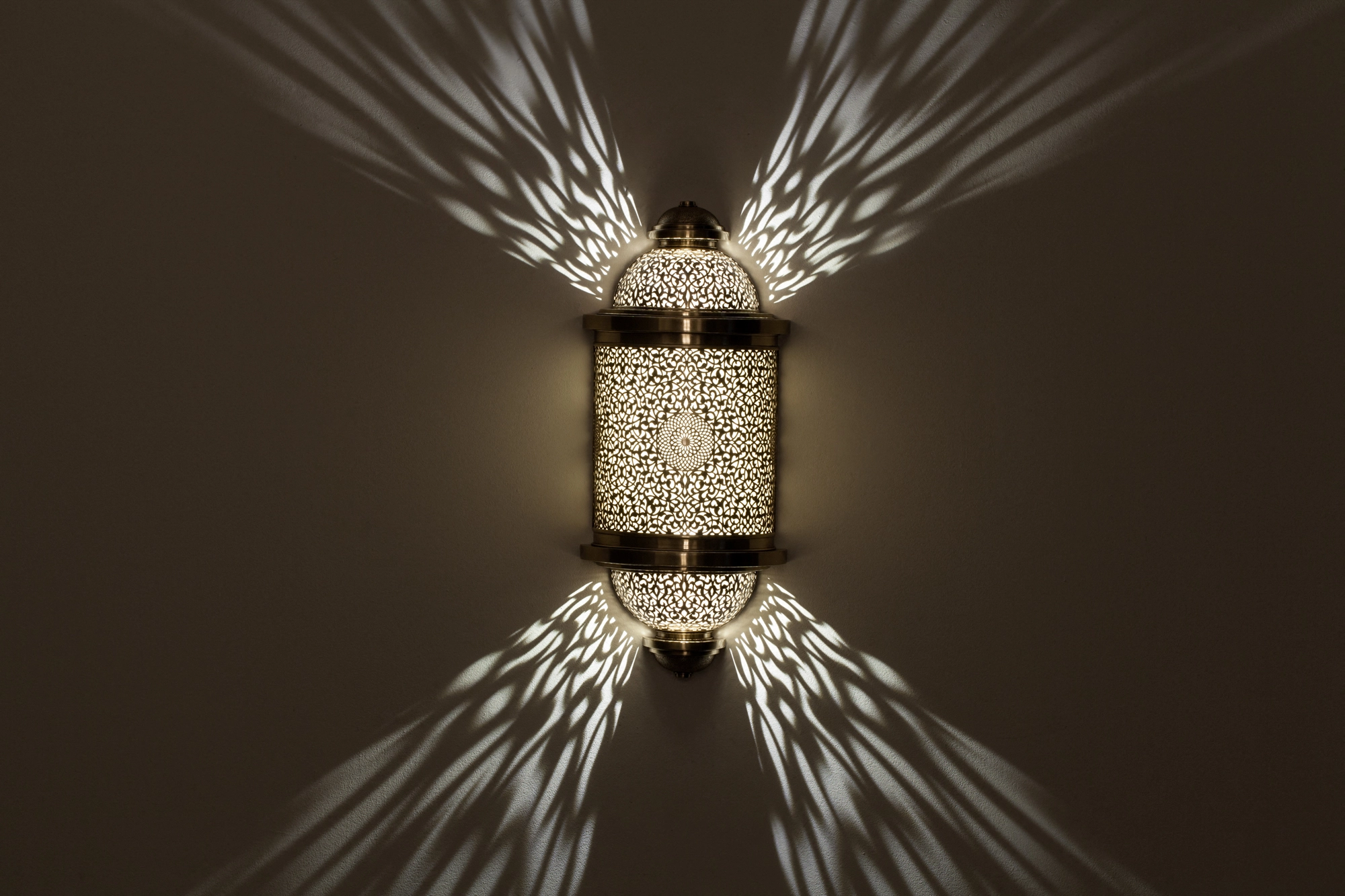 Pierced-brass Treville barrel wall light glowing, casting symmetrical fans of light up and down a dark wall.
