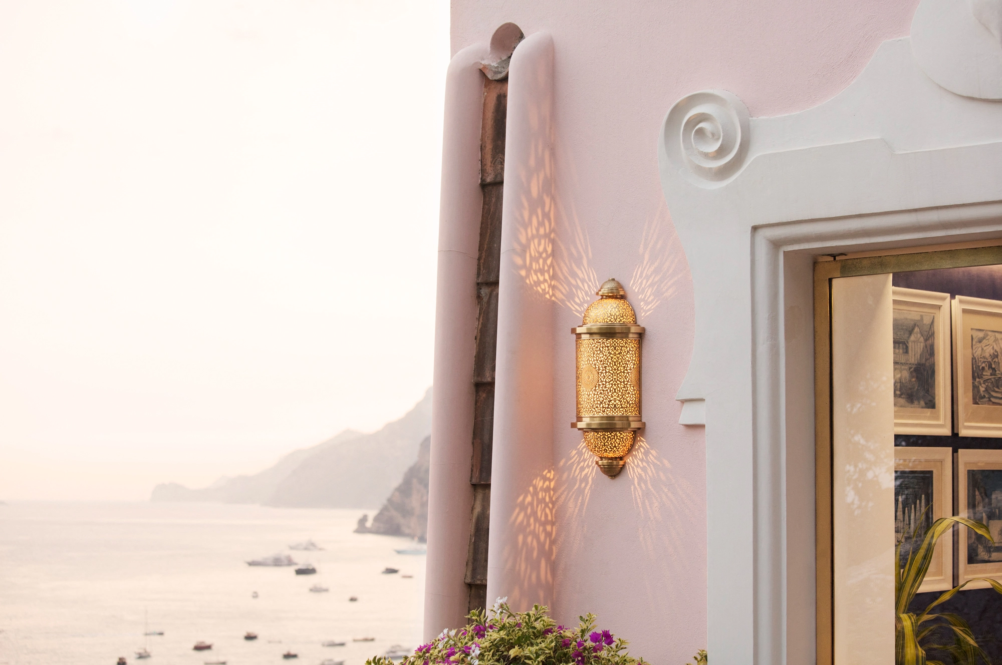 Pink villa wall with a brass lantern beside a window above a hazy coastal view.