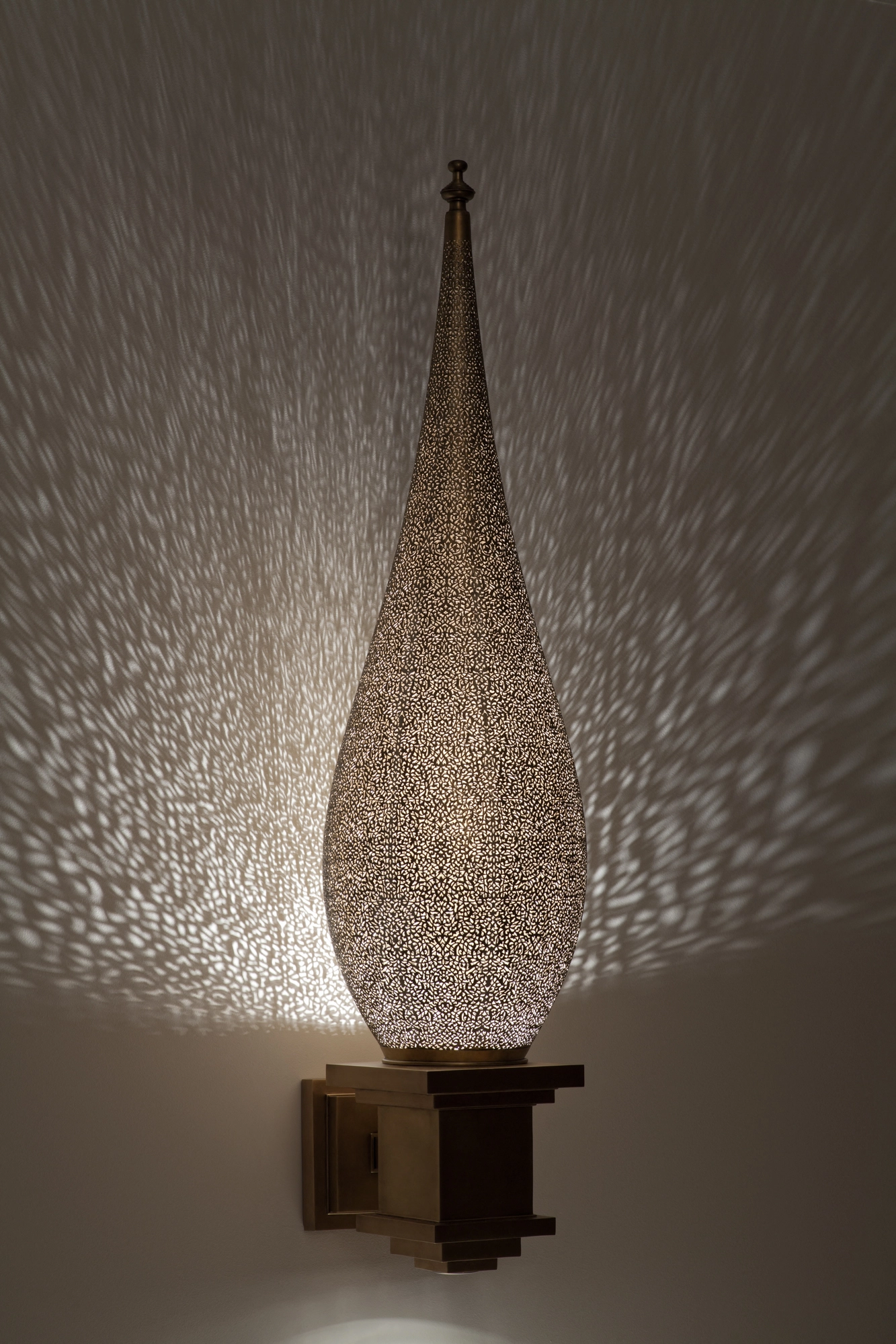Tall pierced-brass seated-flame wall light on a bracket, its teardrop form glowing amid a dense halo of light dots.
