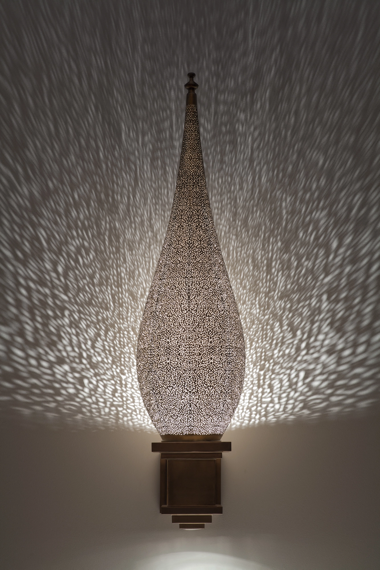 Pierced-brass seated-flame wall light glowing on a bracket, casting a wide halo of fine light dots on the wall.