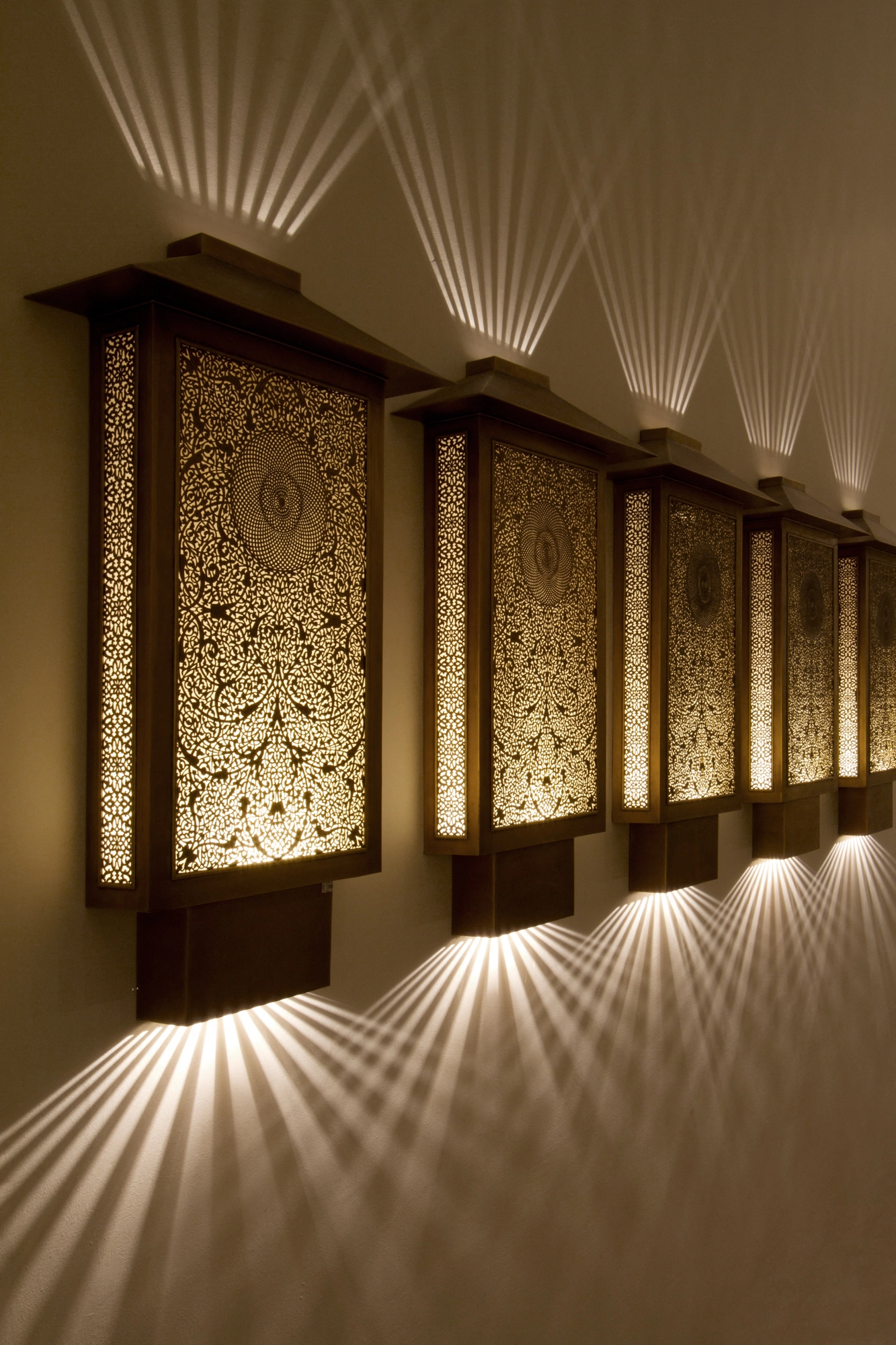 Row of square pierced-brass oriental box wall lights along a corridor, each casting fans of light on the wall.