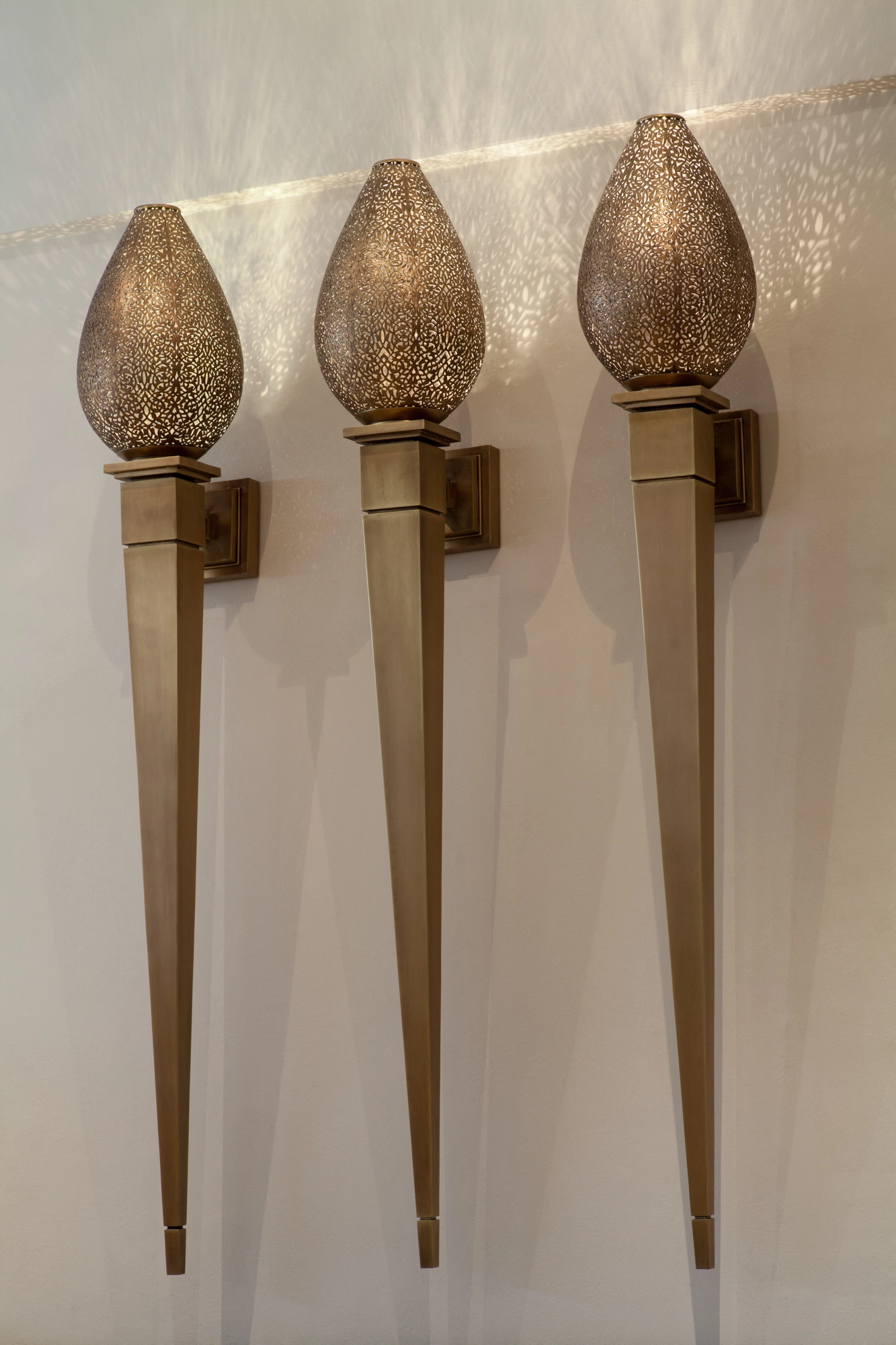 Three pierced-brass lotus-torch wall lights with teardrop tops on tapering stems, mounted in a row.