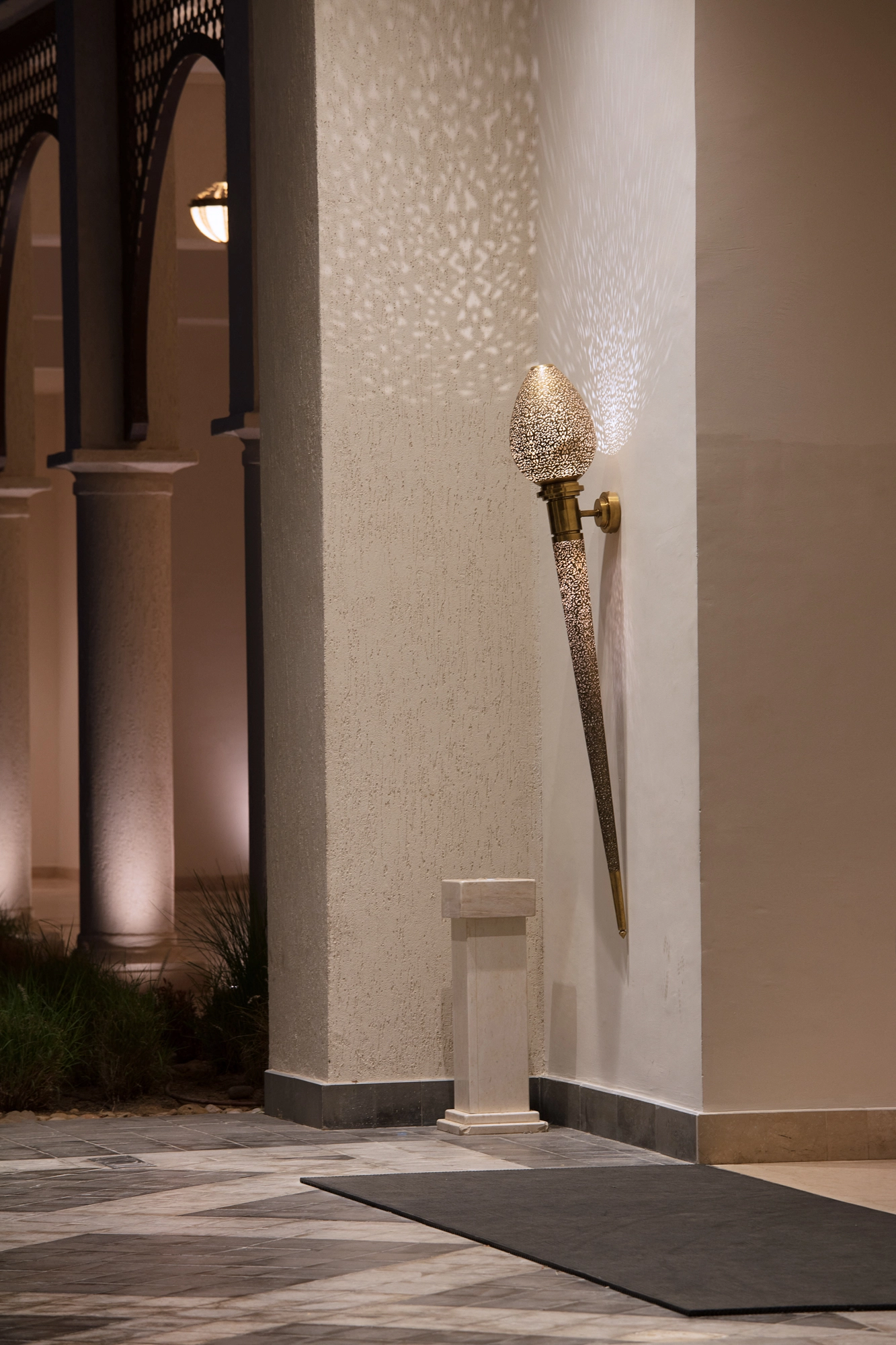 Tall torch-shaped pierced wall light glowing beside a stone column at a paved entrance at dusk.