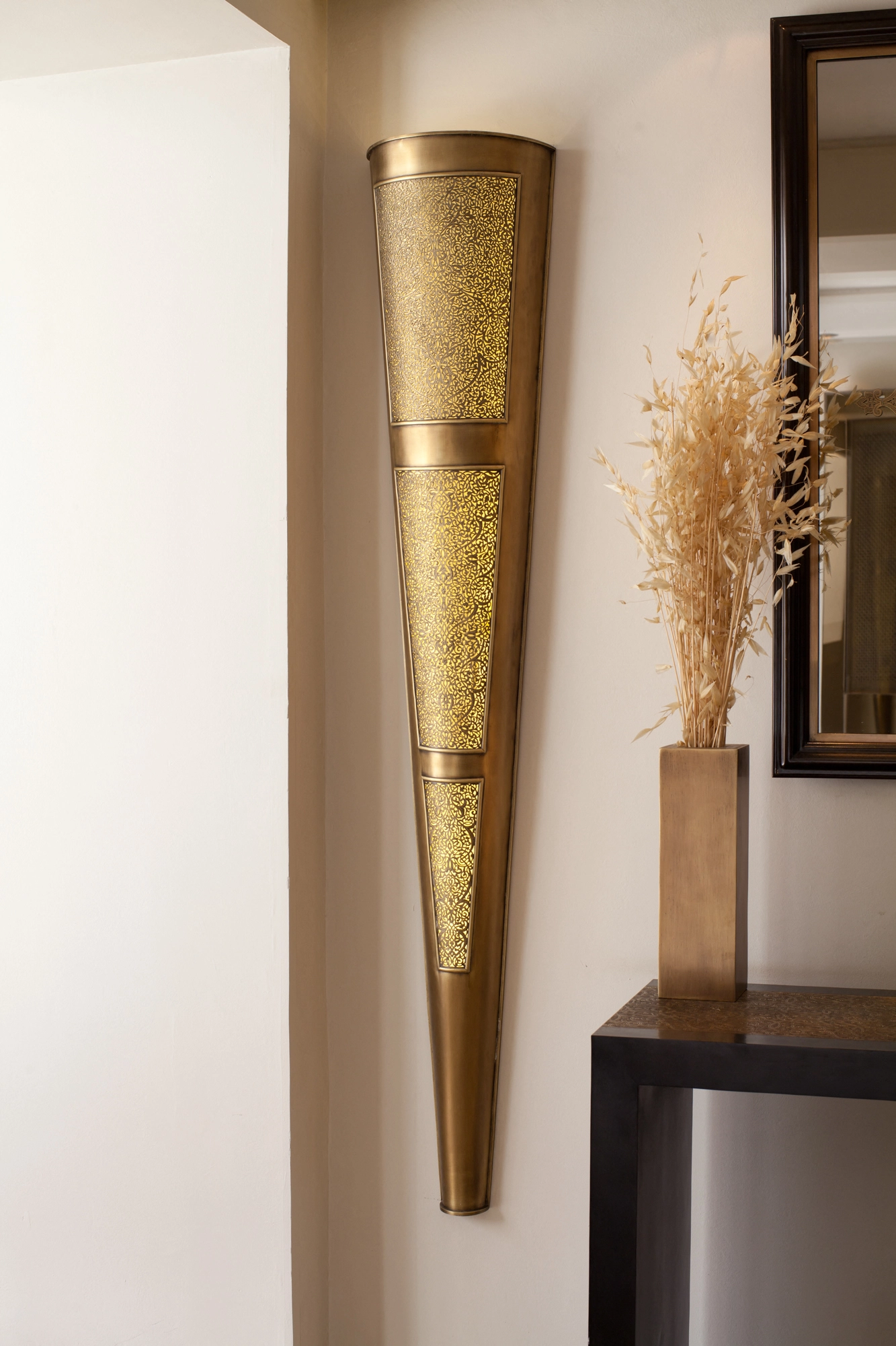 Tall tapering brass half-torch wall light mounted beside a console table with a vase of dried grasses.