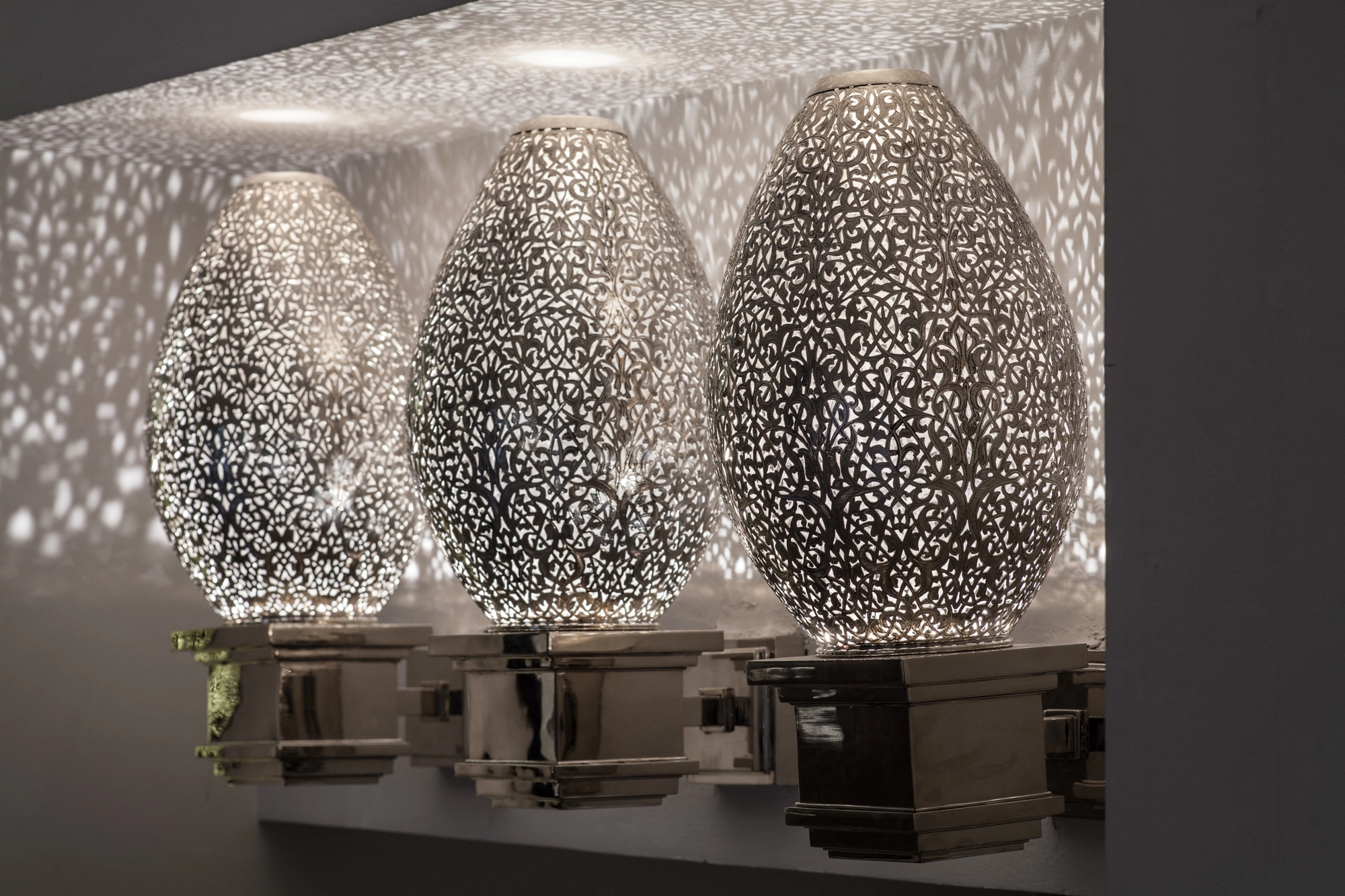 Three pierced-brass seated-amphora wall lights in a row, glowing egg-shaped forms scattering light dots on the wall.