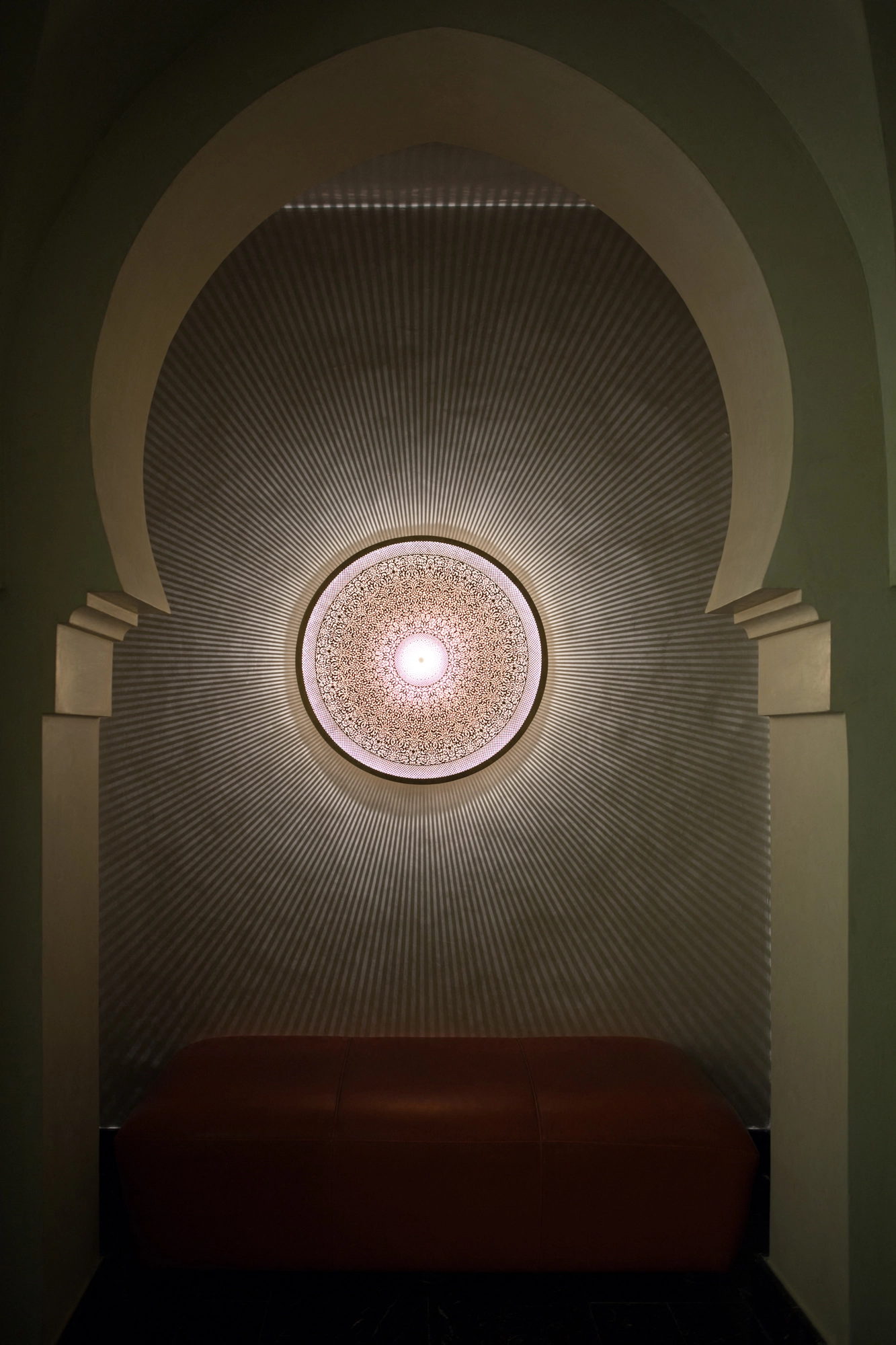 Glowing circular medallion wall light in an arched niche above a red bench.