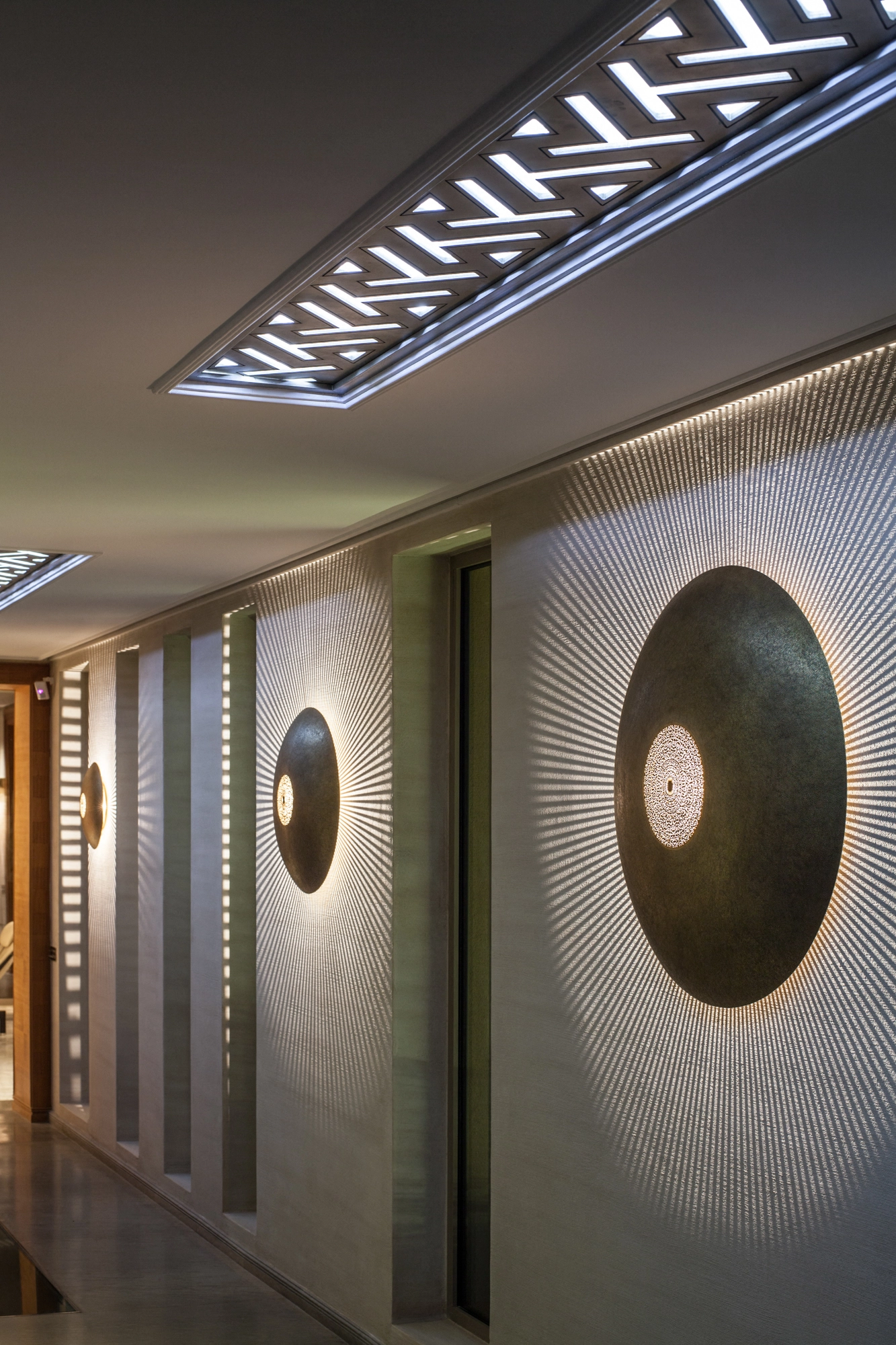 Round pierced-brass shield wall lights mounted along a corridor, each glowing at the center with radiating light.