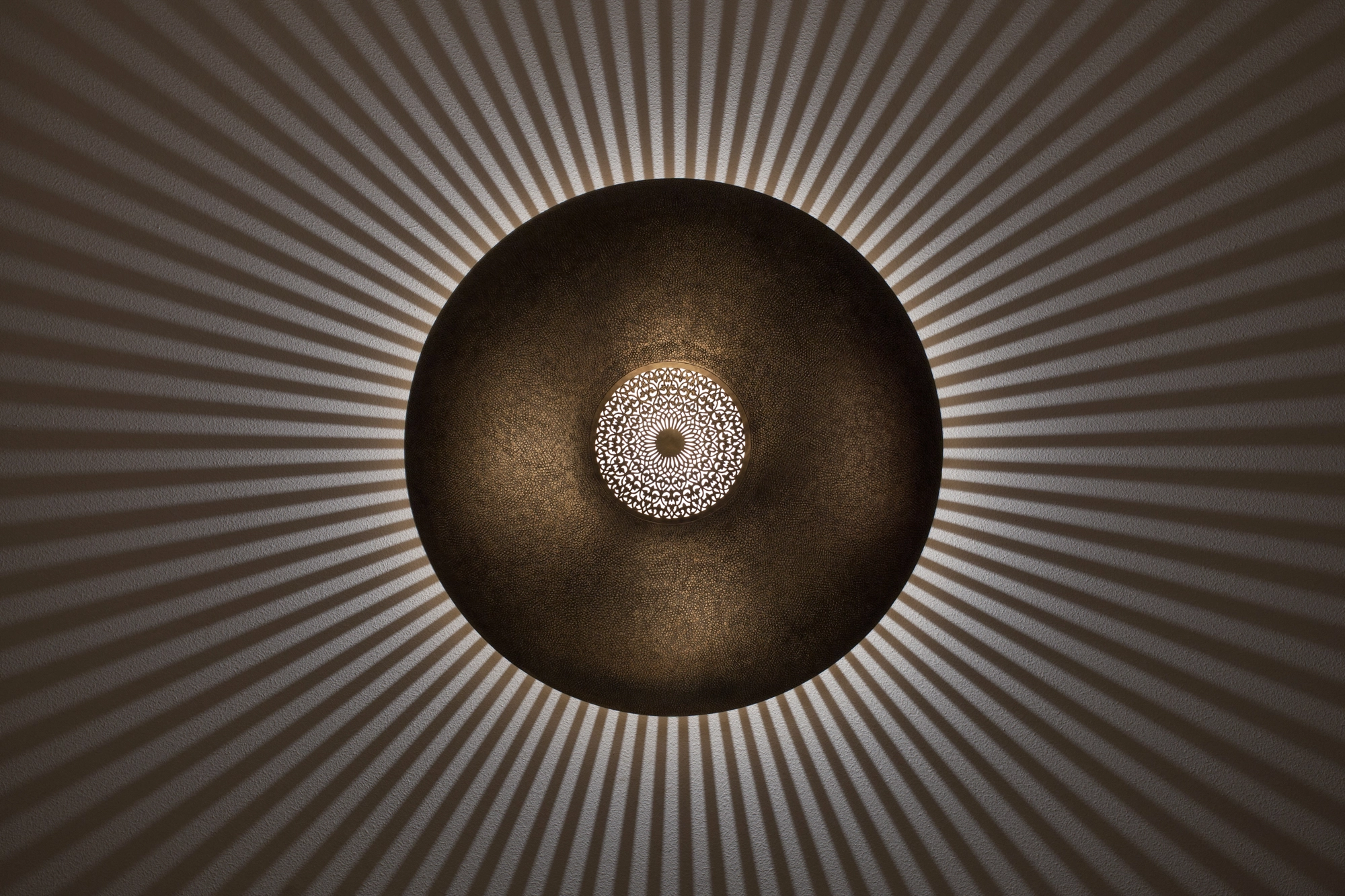 A round pierced metal wall light with a glowing central disc radiating fine dappled light on a dark background.