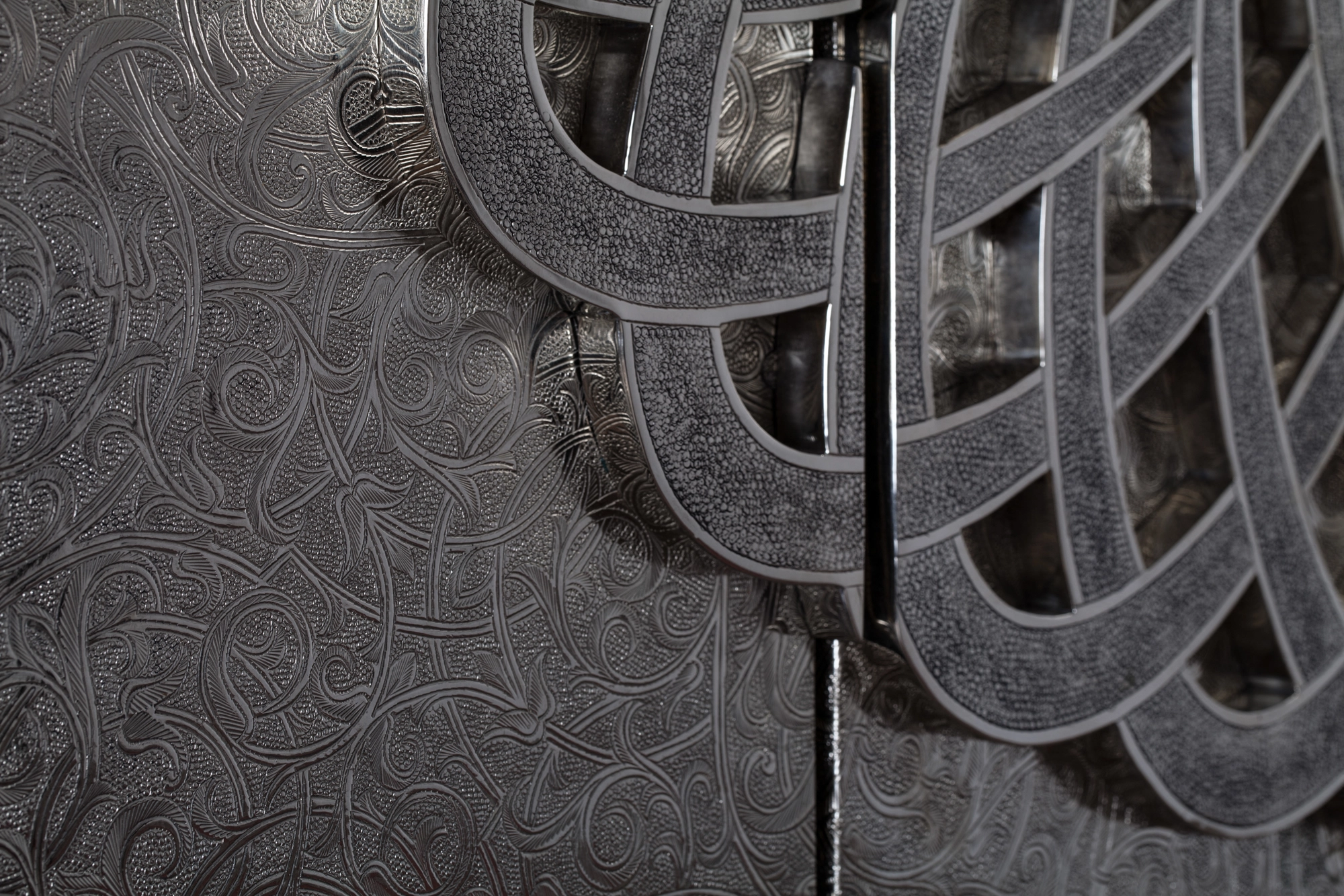 Angled close-up of interwoven silver bands over a finely engraved panel on a wall artwork.