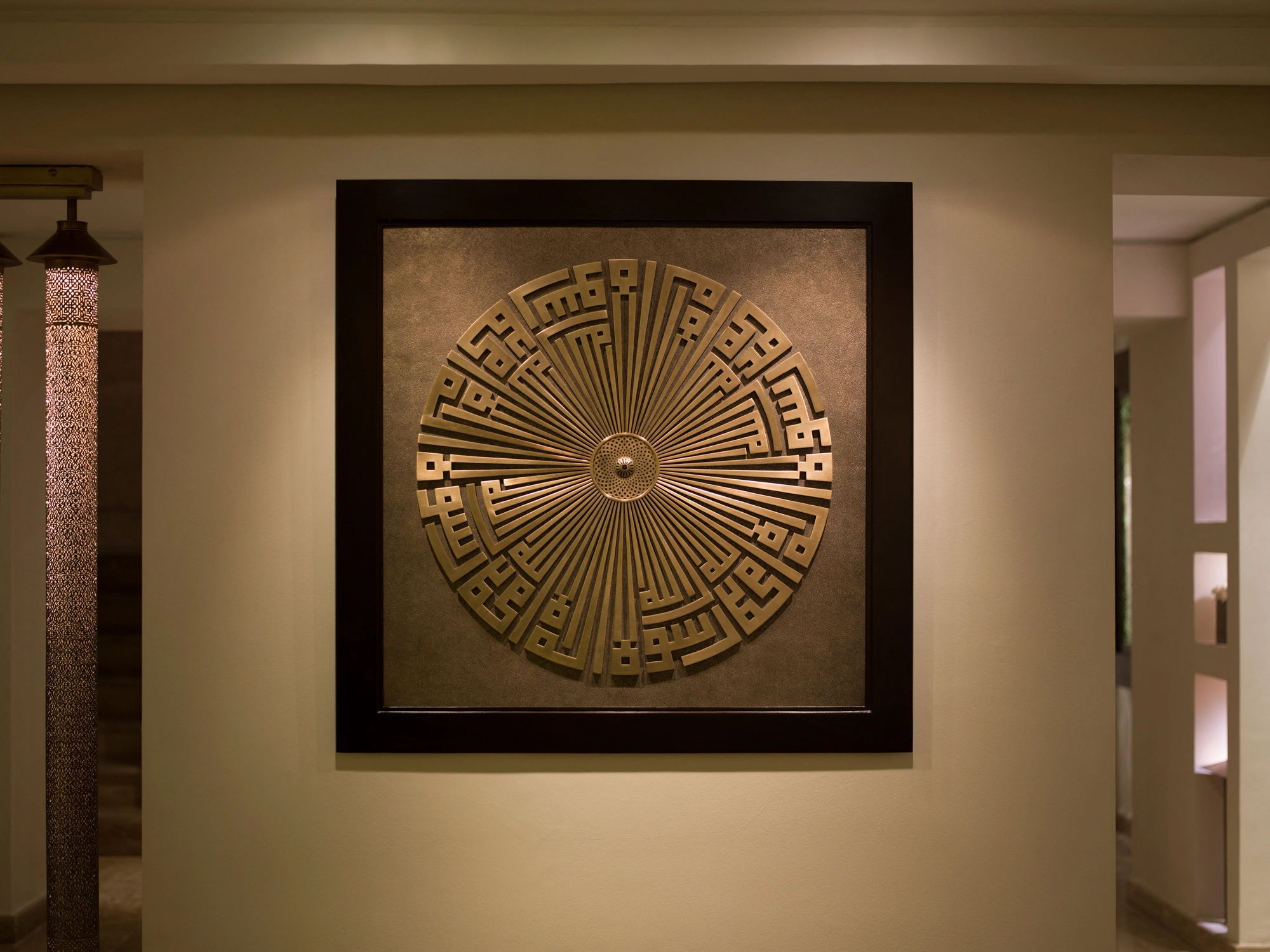 A framed square Kufic medallion artwork mounted on a wall in a warmly lit hallway.