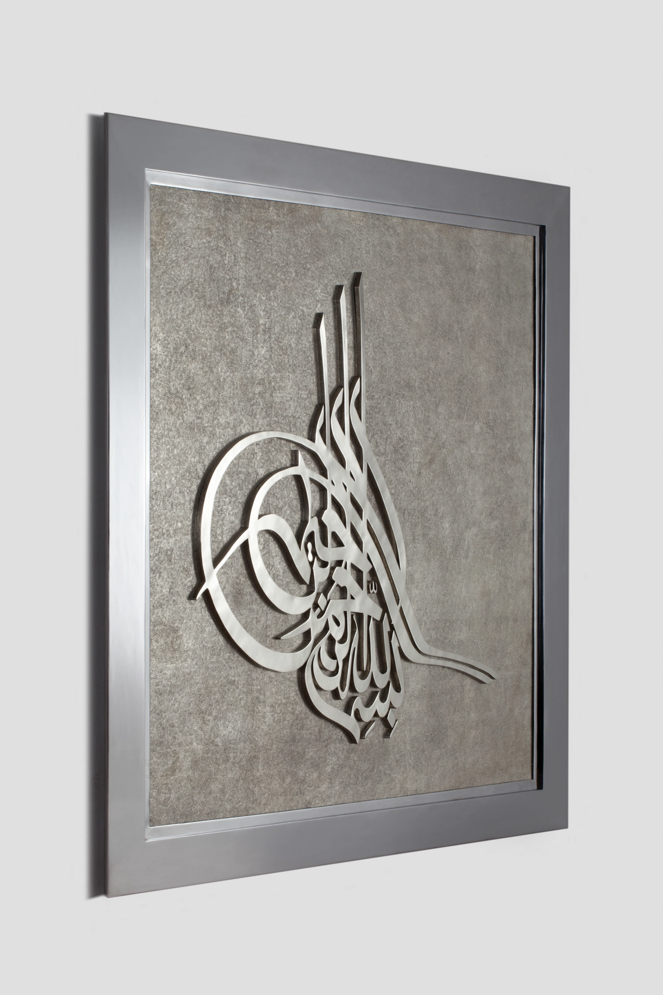 A framed wall panel with a white calligraphic motif on a textured ground, seen at an angle in a grey frame.
