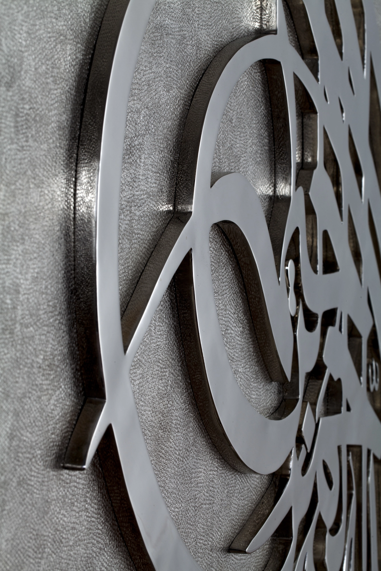 Close-up of curved white metal calligraphic strokes raised over a textured grey panel.