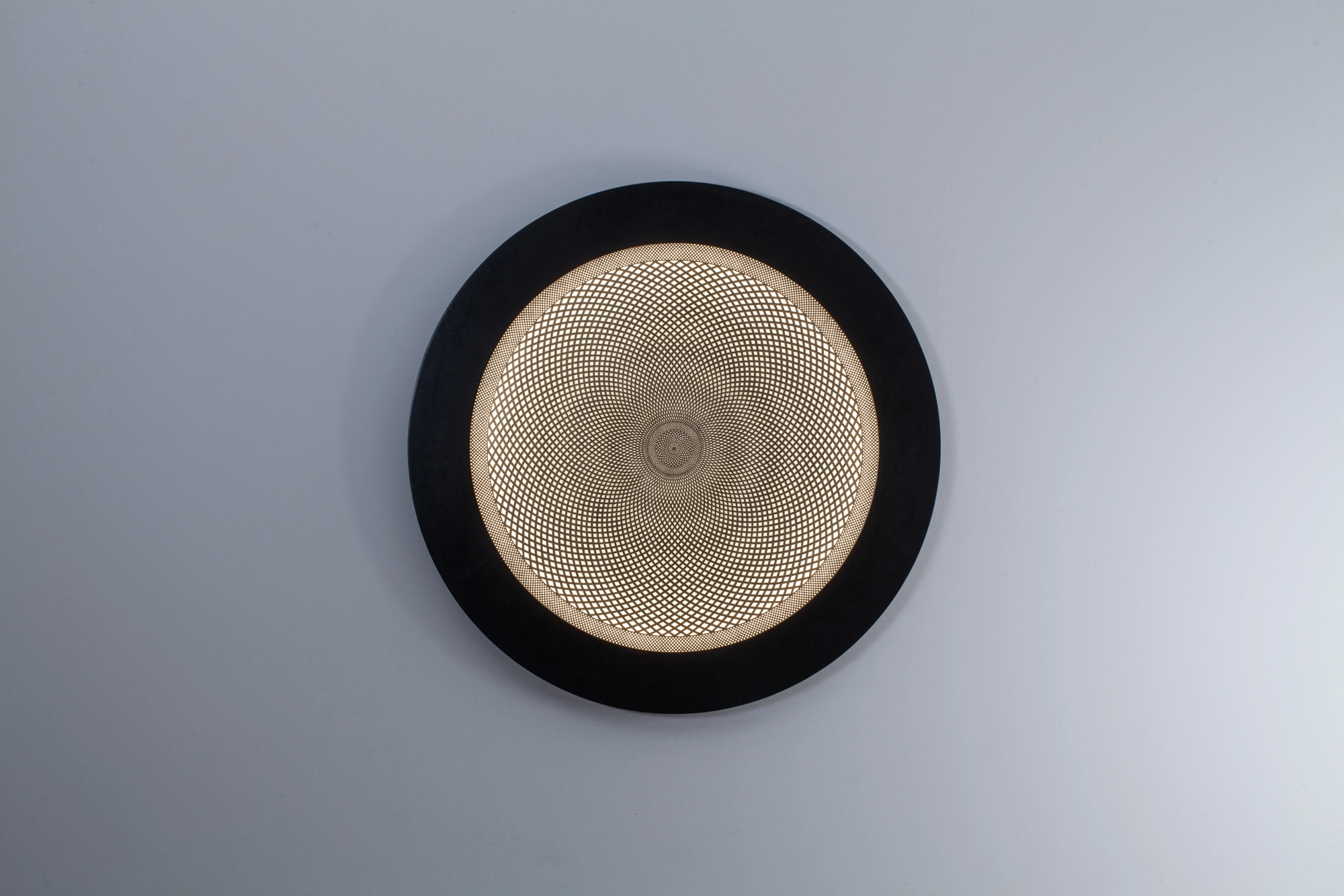 A round backlit wall disc with a perforated golden center in a black ring frame, viewed at an angle on a pale wall.