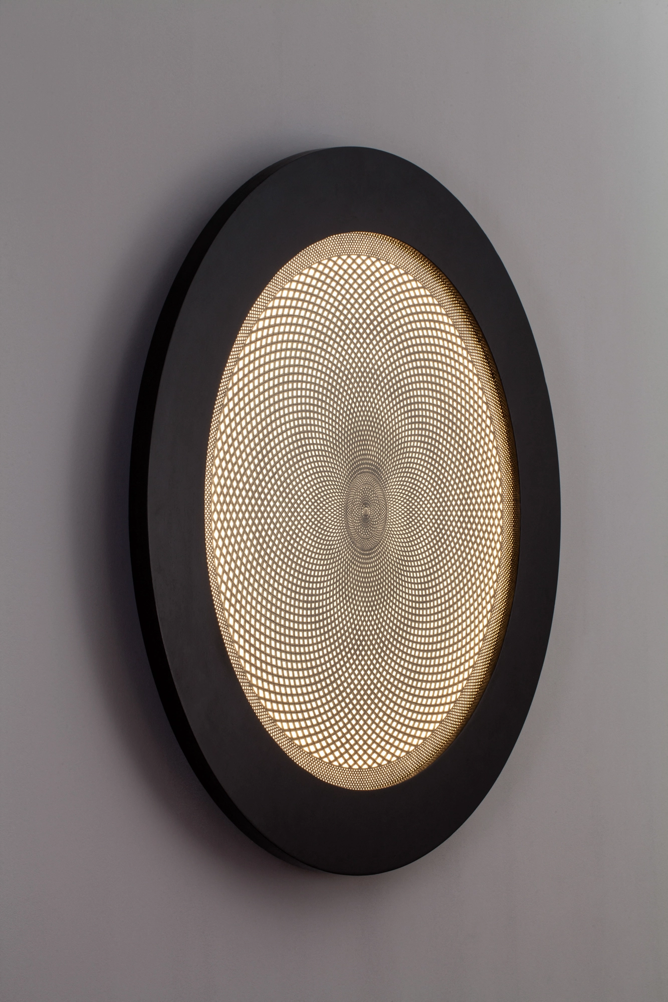 An oval backlit wall disc with a finely perforated golden center in a thick black ring frame on a grey wall.