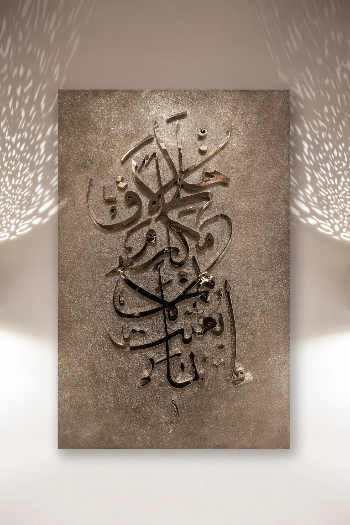A tall wall panel of raised metallic Arabic calligraphy on a mottled earth-toned ground.