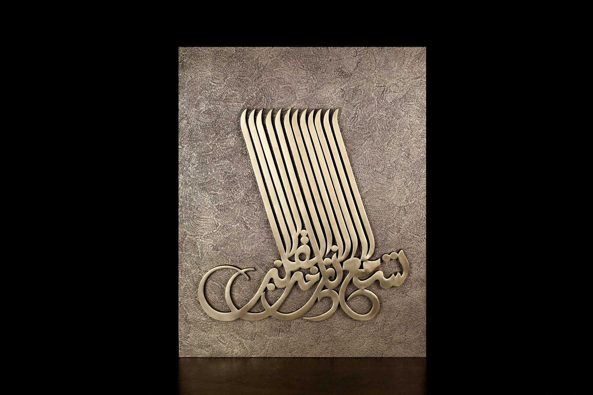 A silver wall panel bearing raised flowing Arabic calligraphy, lit against a black background.