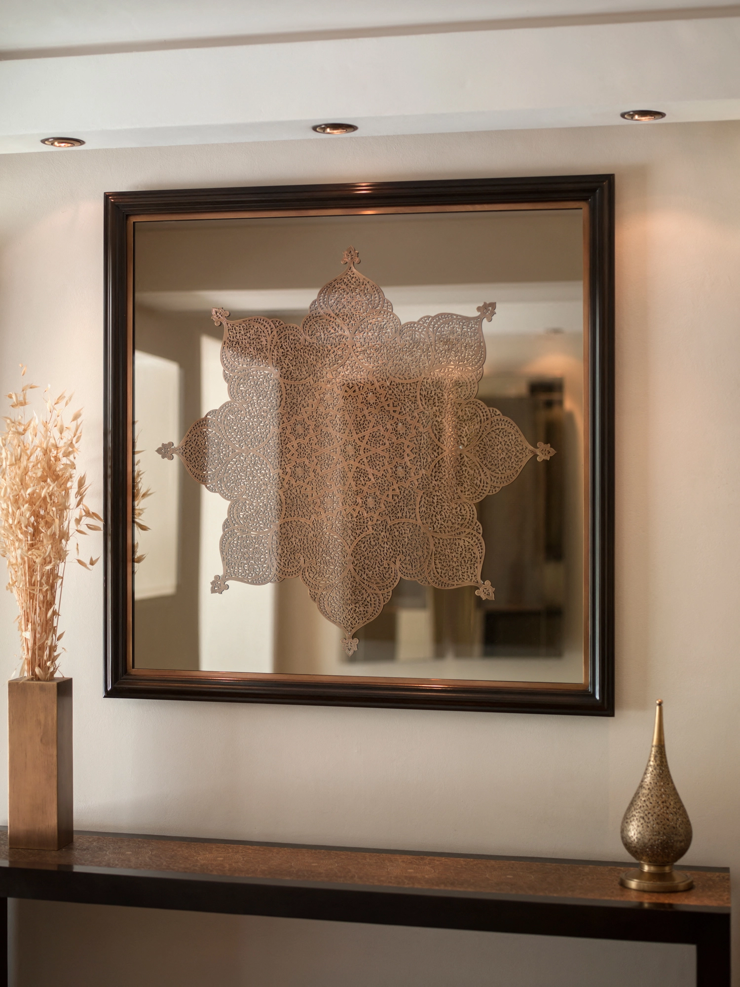 A star-shaped pierced silver wall panel reflected in a framed mirror above a console with a brass vase.