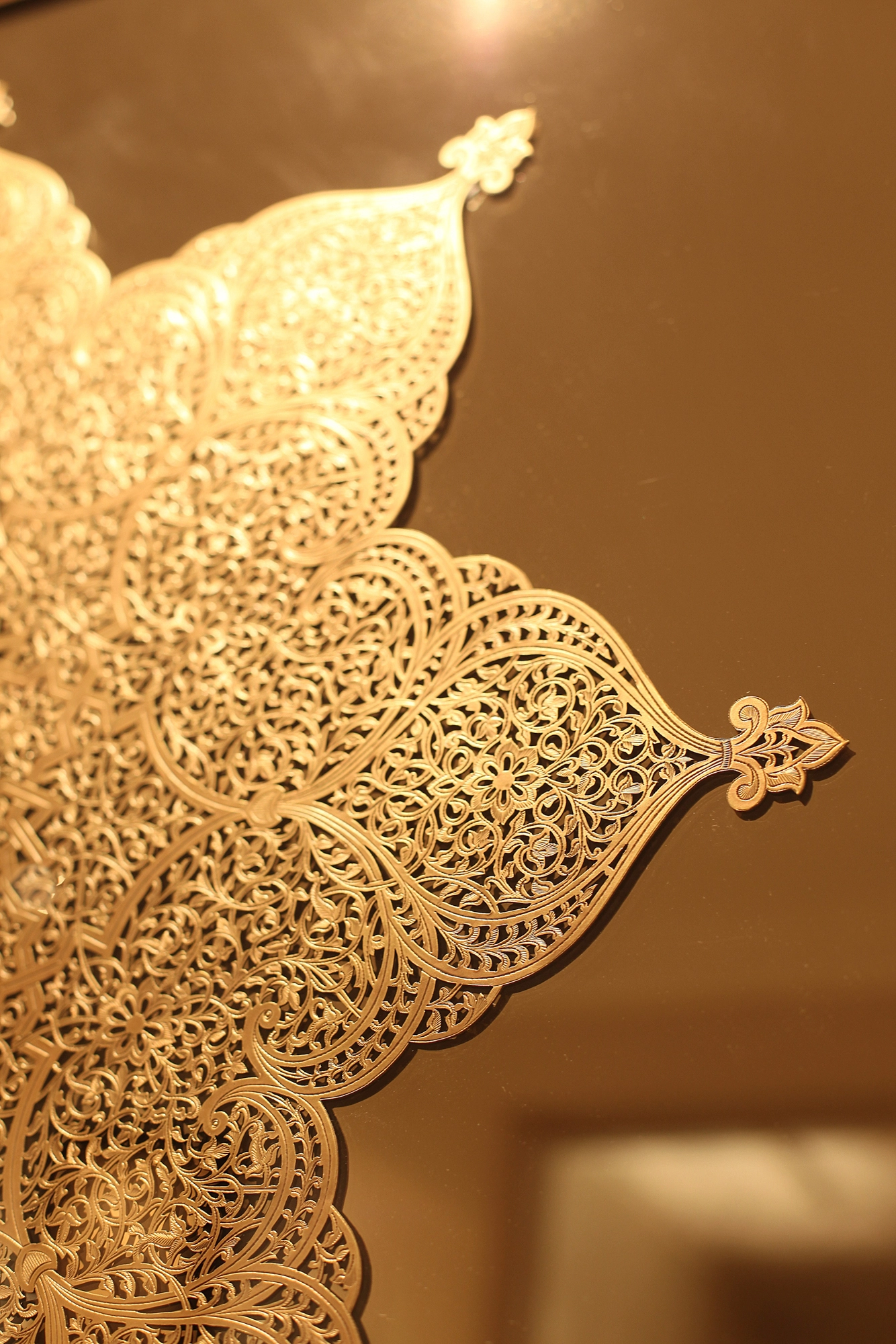 Close-up of a golden pierced filigree panel in a teardrop shape, glowing warmly against a blurred interior.