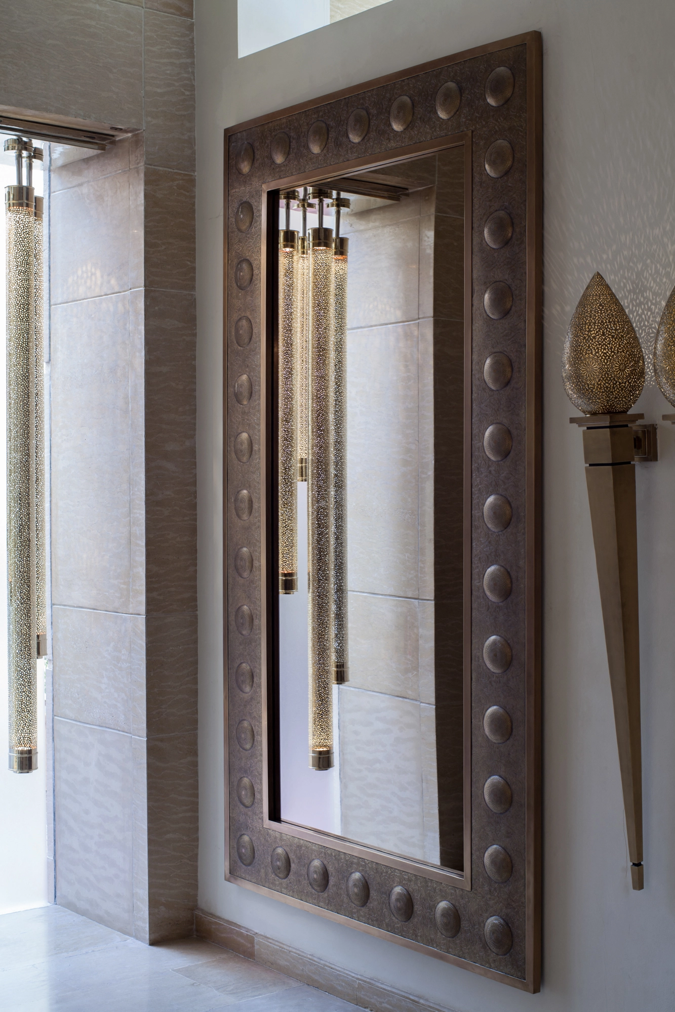 A tall rectangular mirror in a studded bronze frame, flanked by glass-rod wall lights and a torch sconce.