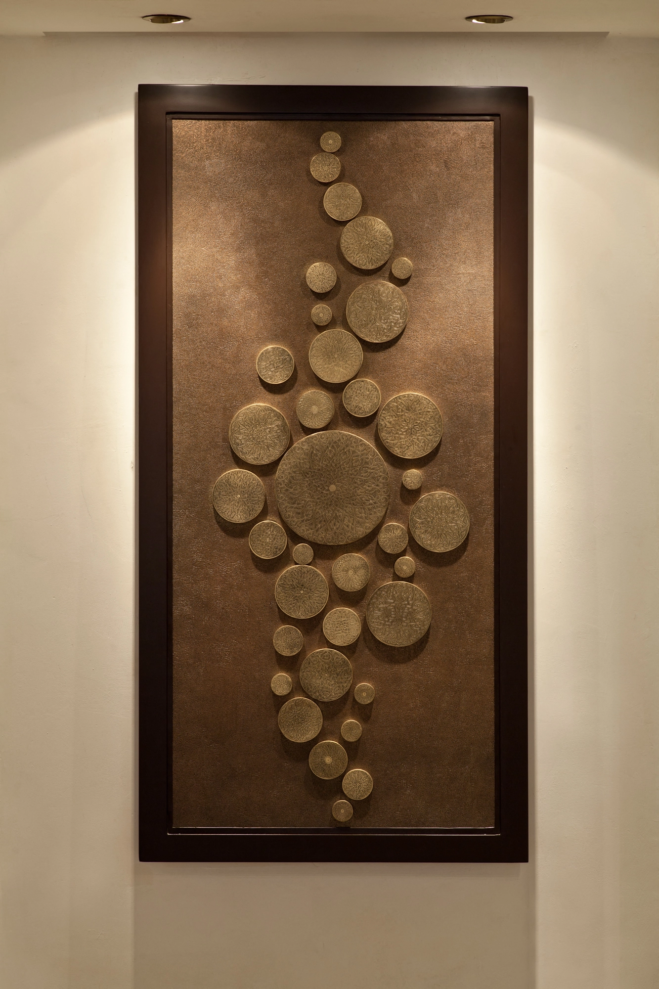 A tall framed wall panel composed of raised bronze discs and circles in varied sizes, on a warm wall.