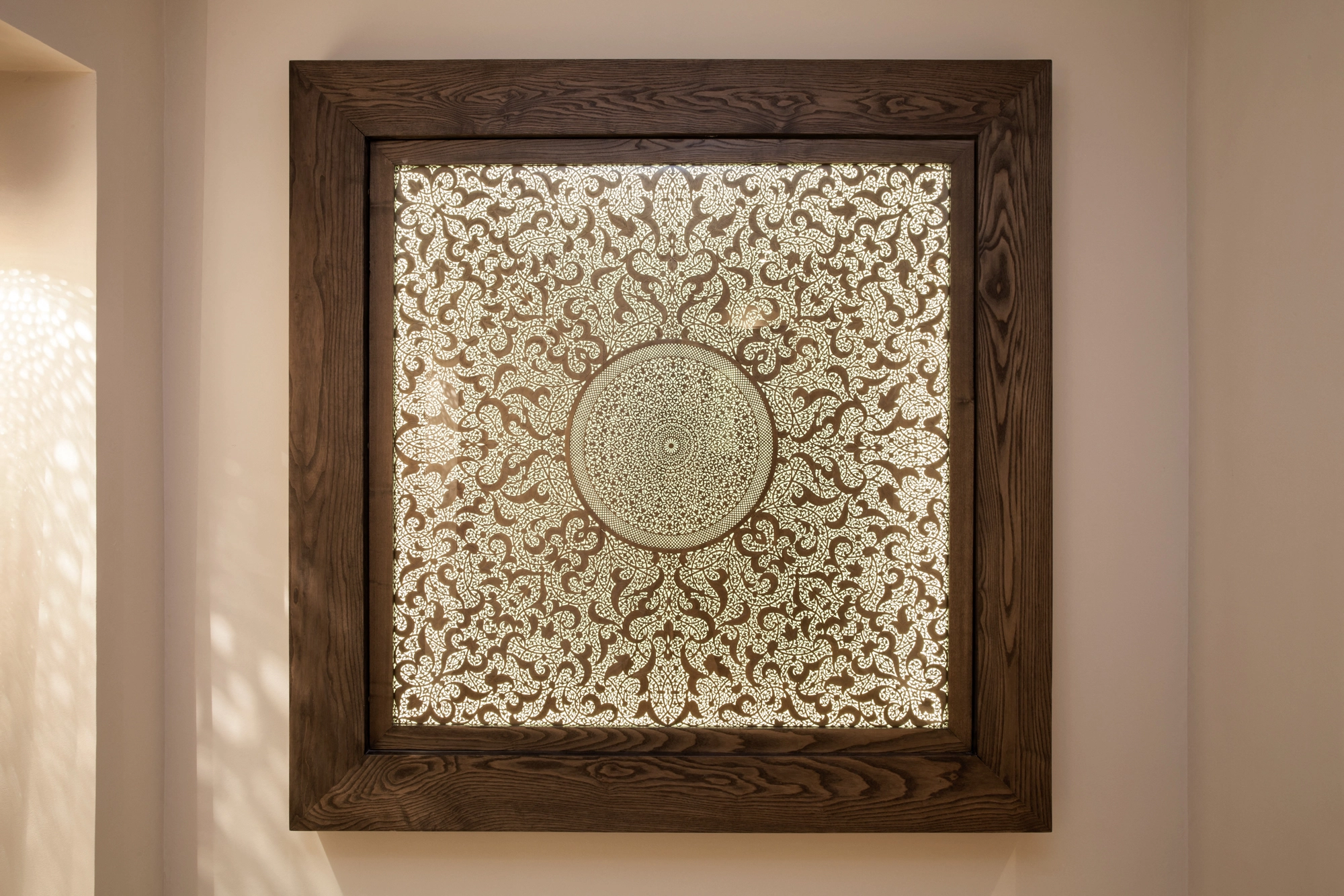 A square backlit wall panel with a pale pierced floral pattern and central medallion in a wooden frame.