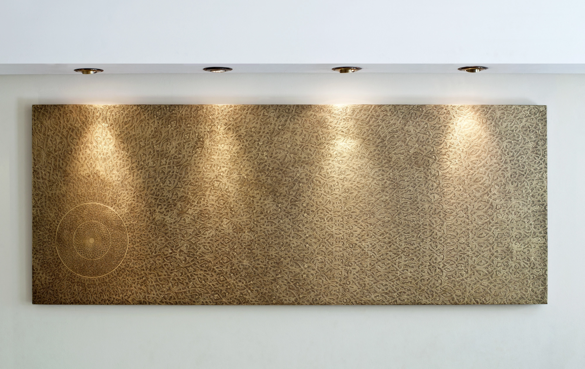 A long horizontal gold textured wall panel lit from above by four downlights against a white wall.