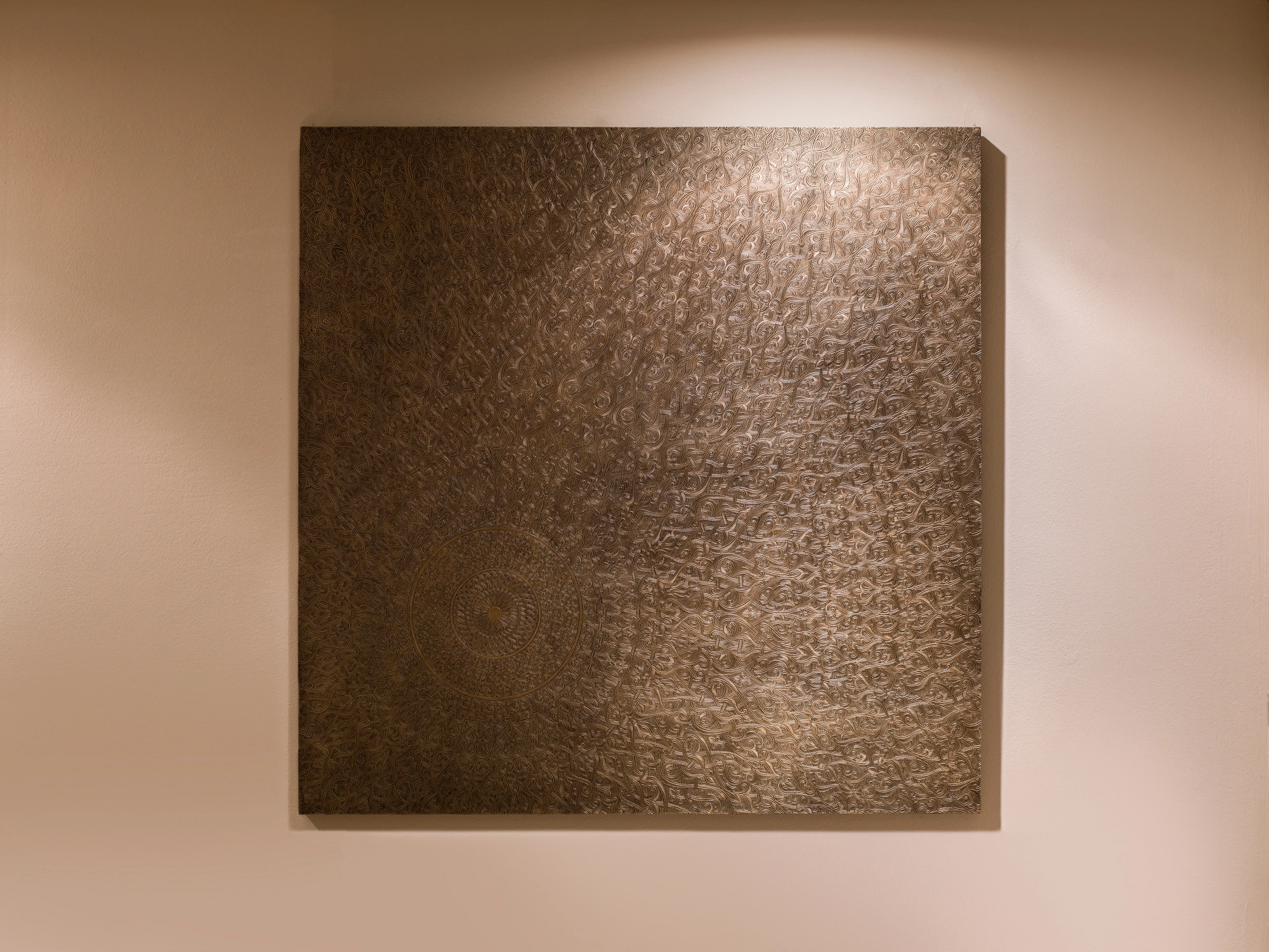 A square textured metal wall panel mounted on a warm wall and lit from one side.