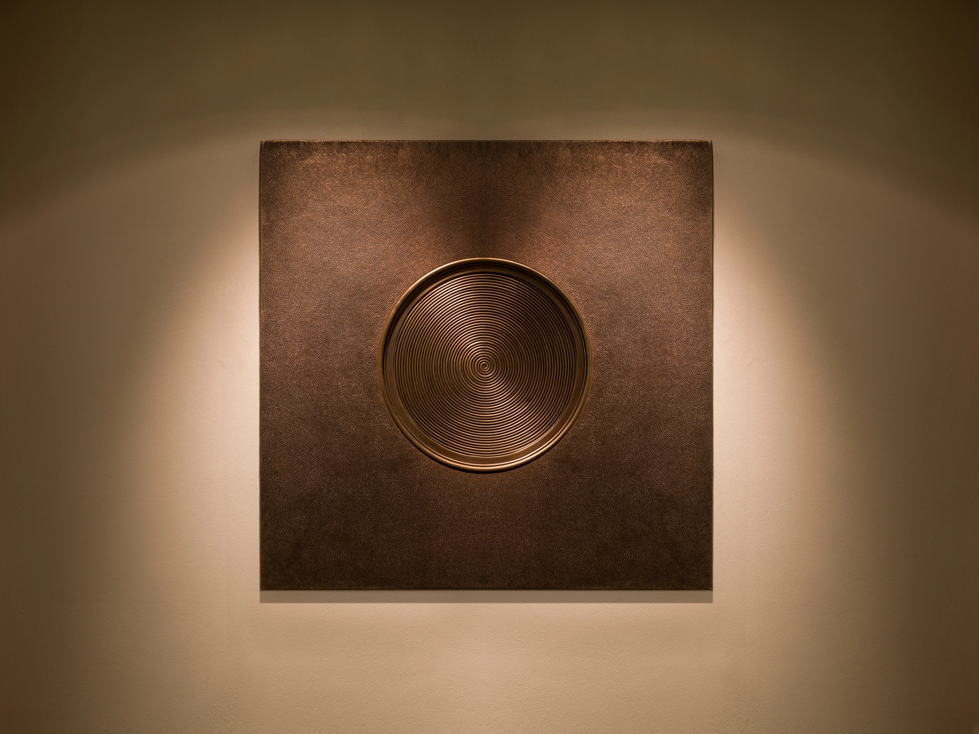 A square wall panel centred on a raised metal disc, warmly spotlit against a pale wall.