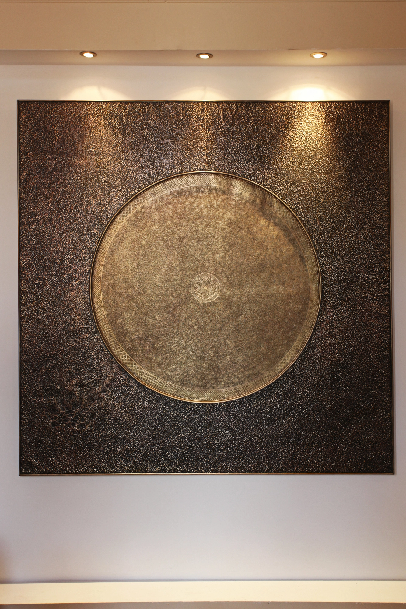 A large dark wall panel centred on an engraved circular metal disc, lit from above.