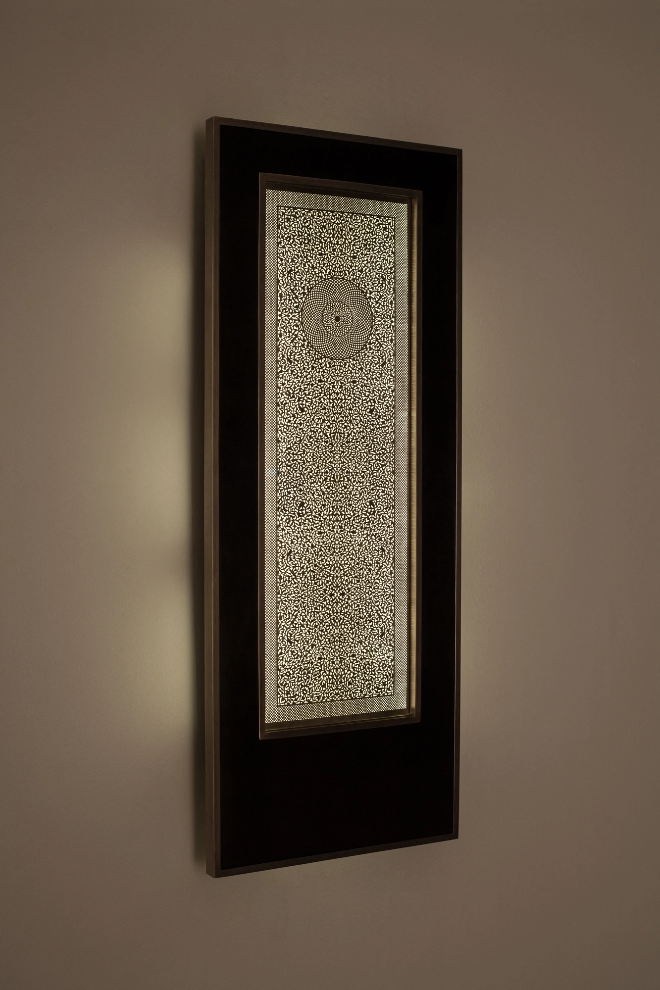 A tall narrow backlit wall panel with intricate pale openwork in a dark frame, glowing against a beige wall.