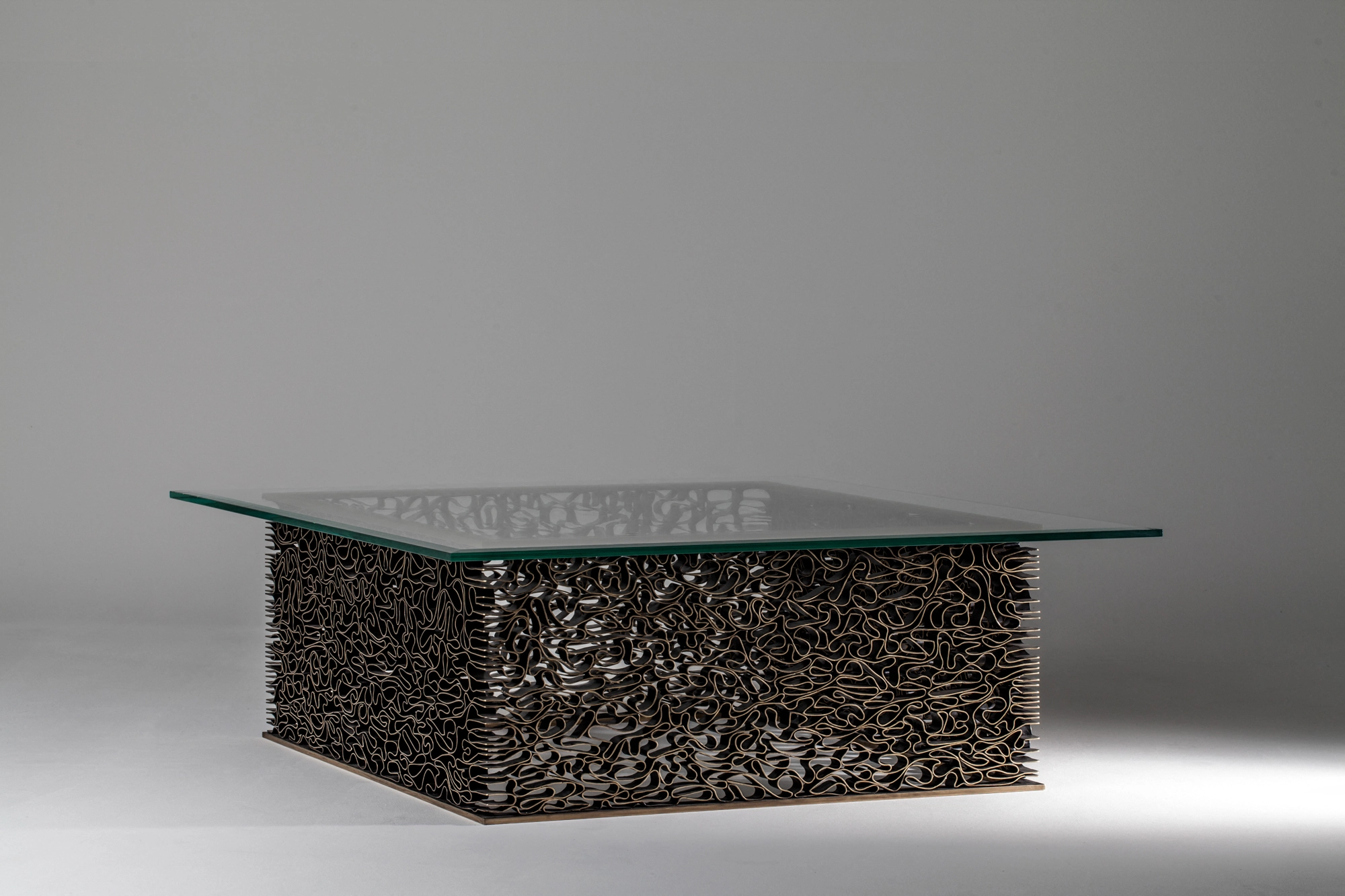 A glass-topped coffee table on a solid block base of densely woven wavy metal, against a grey background.