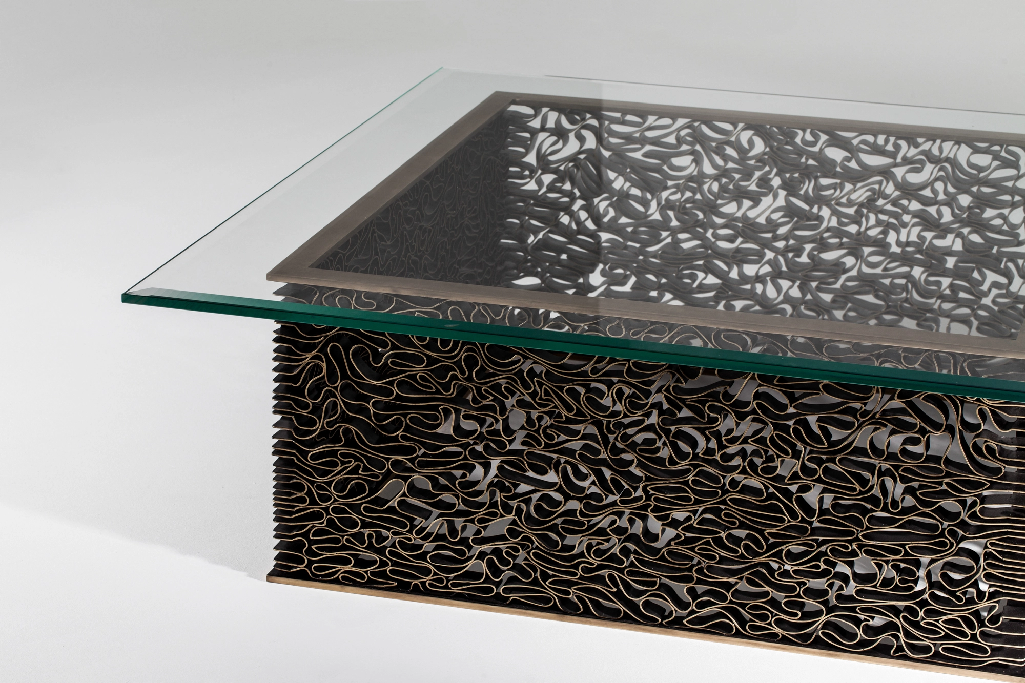 A glass-topped coffee table on a block base of woven wavy metal, viewed from the side on grey.