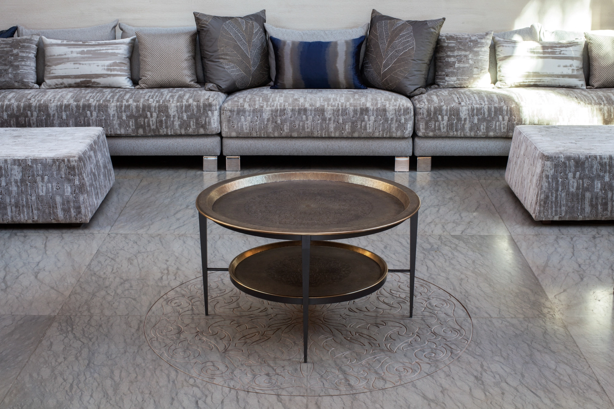 A round two-tier brass tray table standing on a rug before a long sofa in a sitting room.