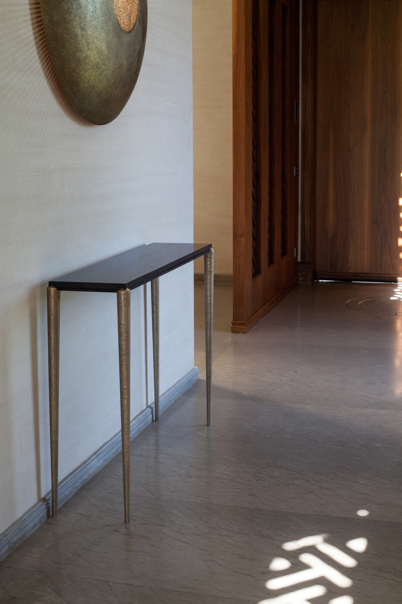 A slim dark console table in a sunlit hallway, with a patterned light cast on the stone floor and a disc above.
