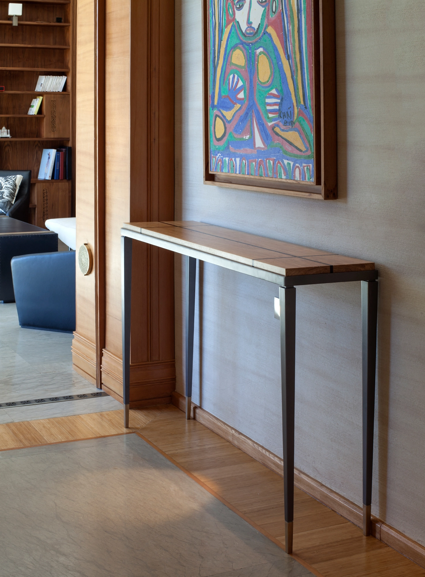 Slim metal console table in a hallway beneath a colourful abstract painting, with a blue bench nearby.
