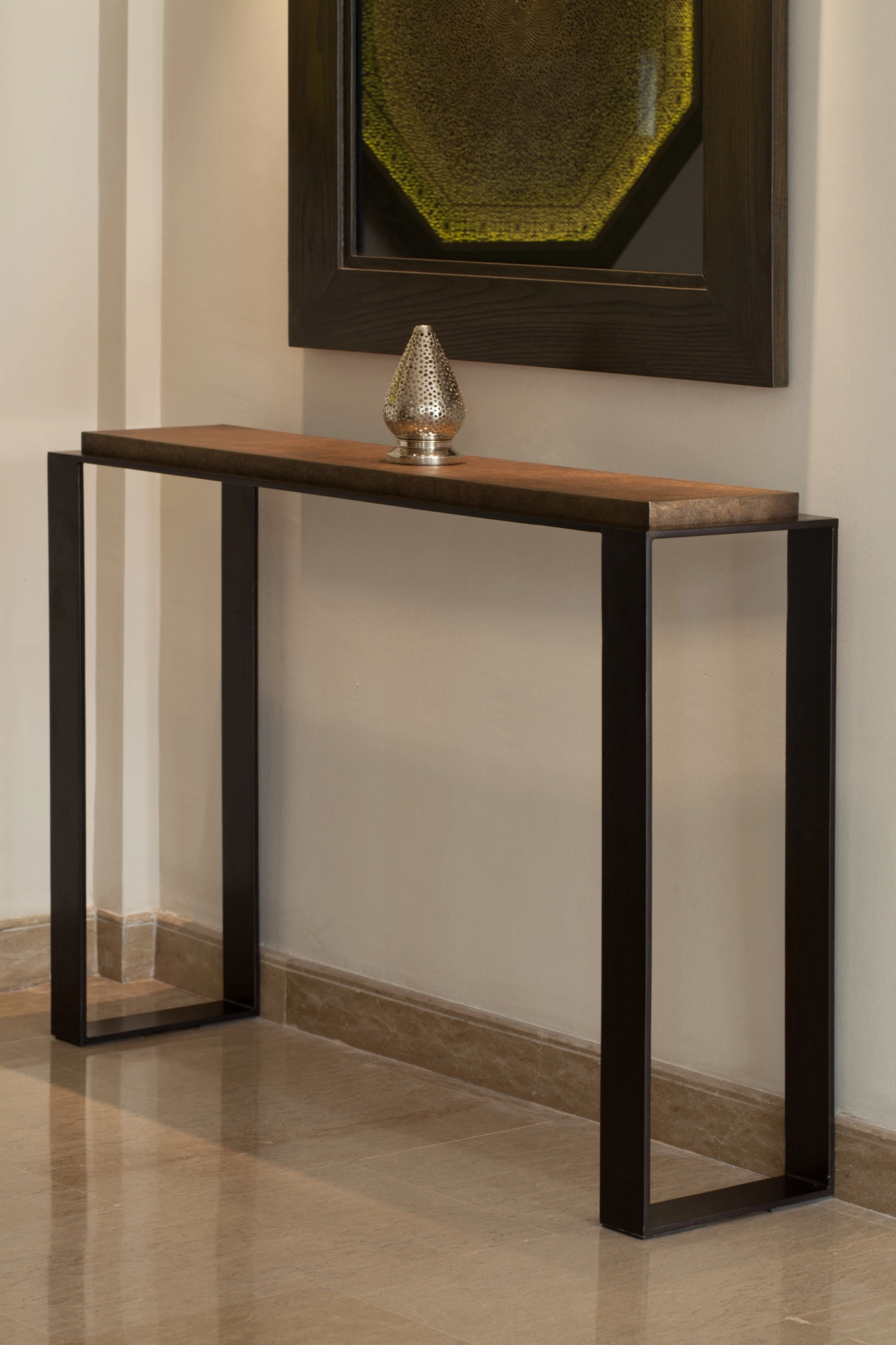 A slim console table with a wood top and dark frame legs beneath a framed artwork, a small ornament on top.