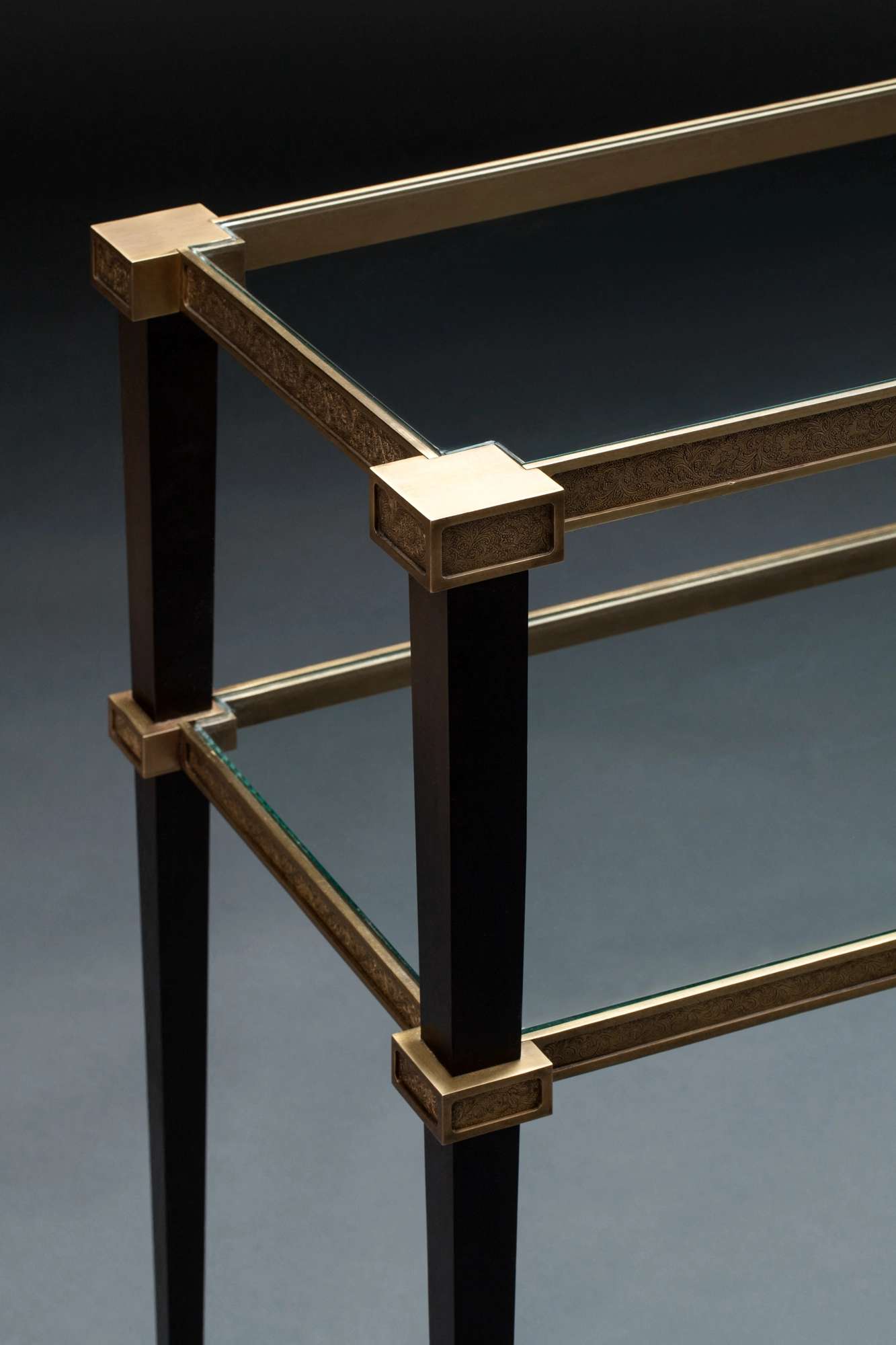 Corner detail of a two-tier glass console table with brass corner mounts, against a grey background.