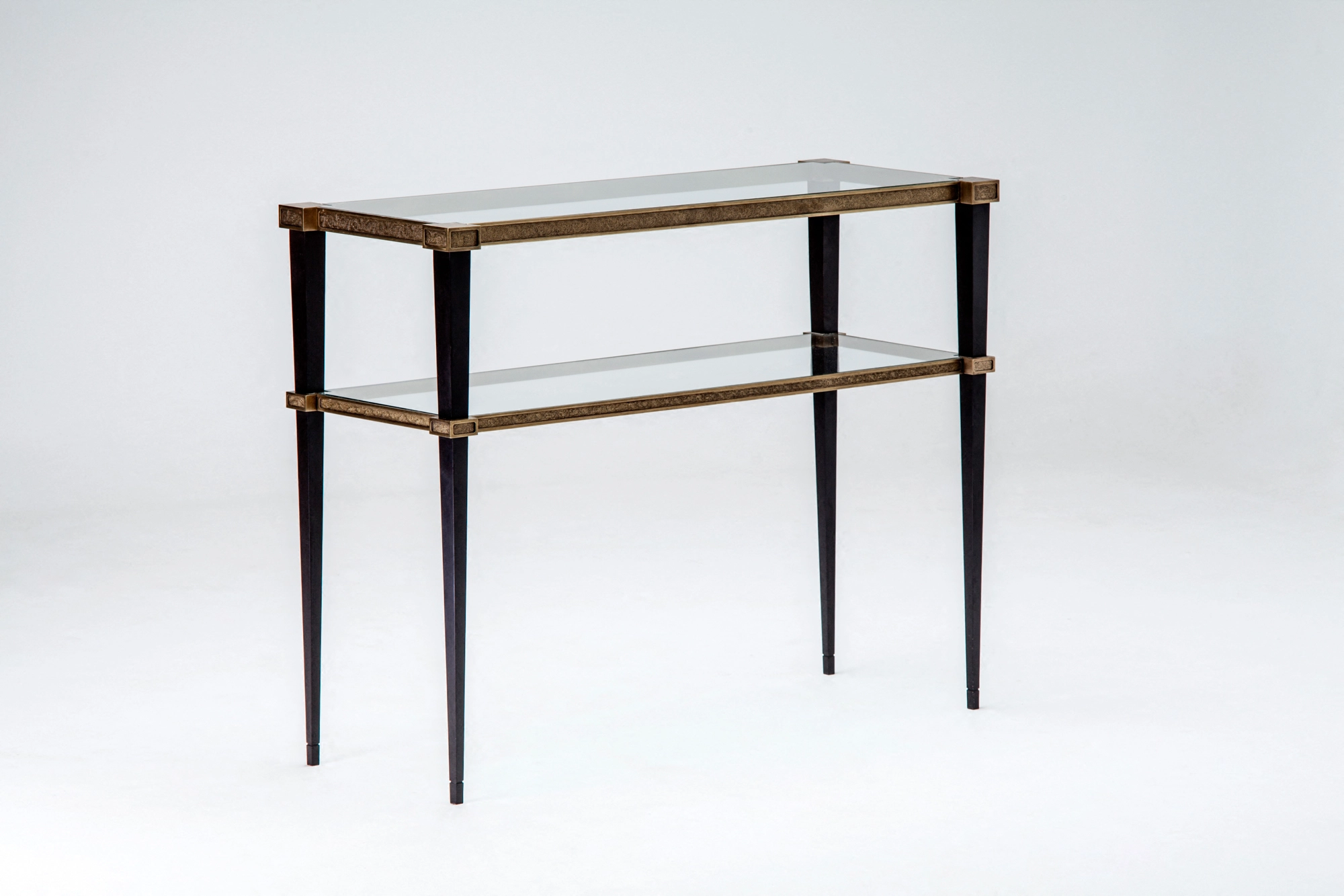 A two-tier glass console table with a brass frame and slim tapered legs, against a white background.