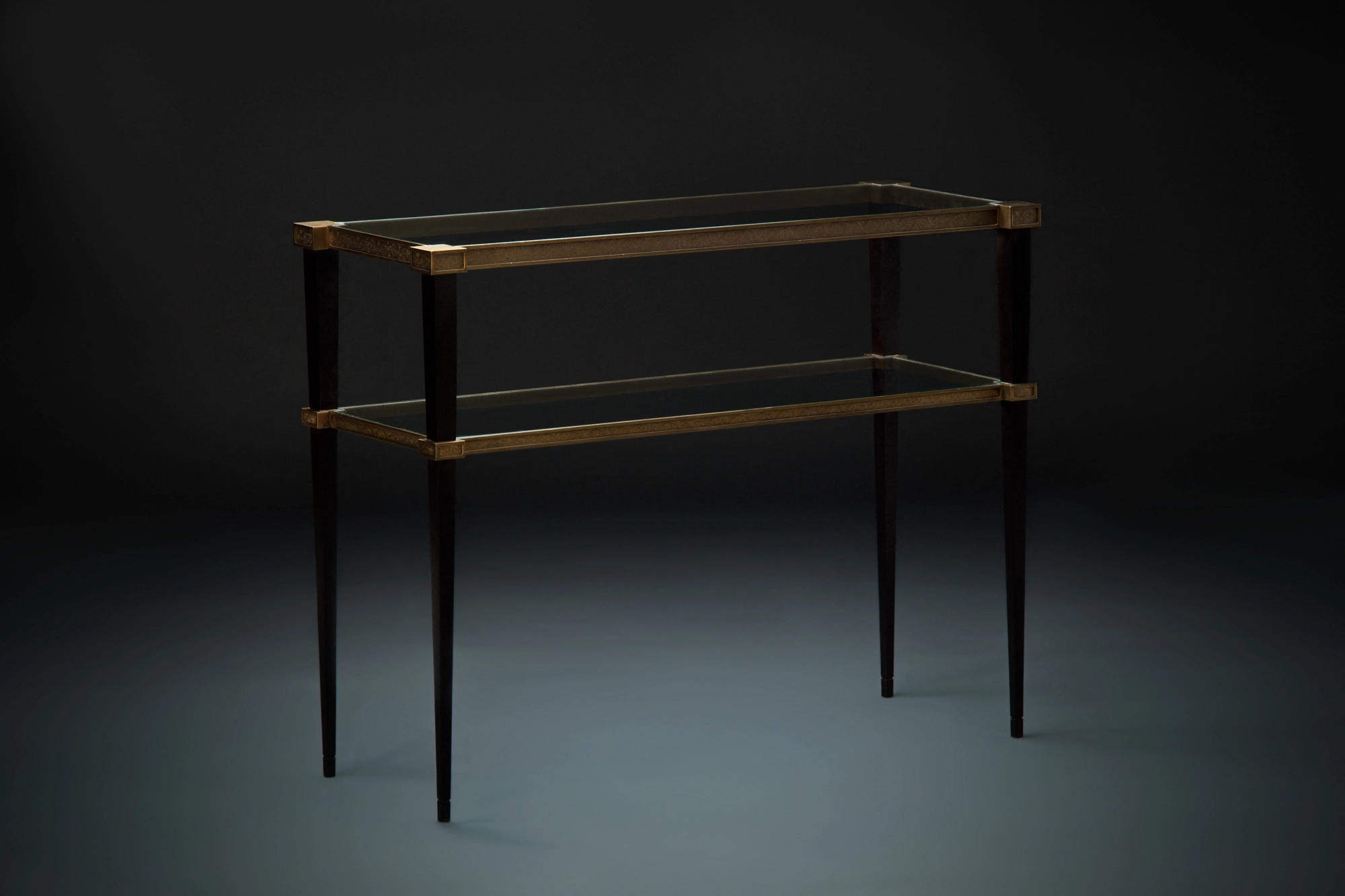 A two-tier glass console table with a brass frame and slim legs, lit against a dark teal background.