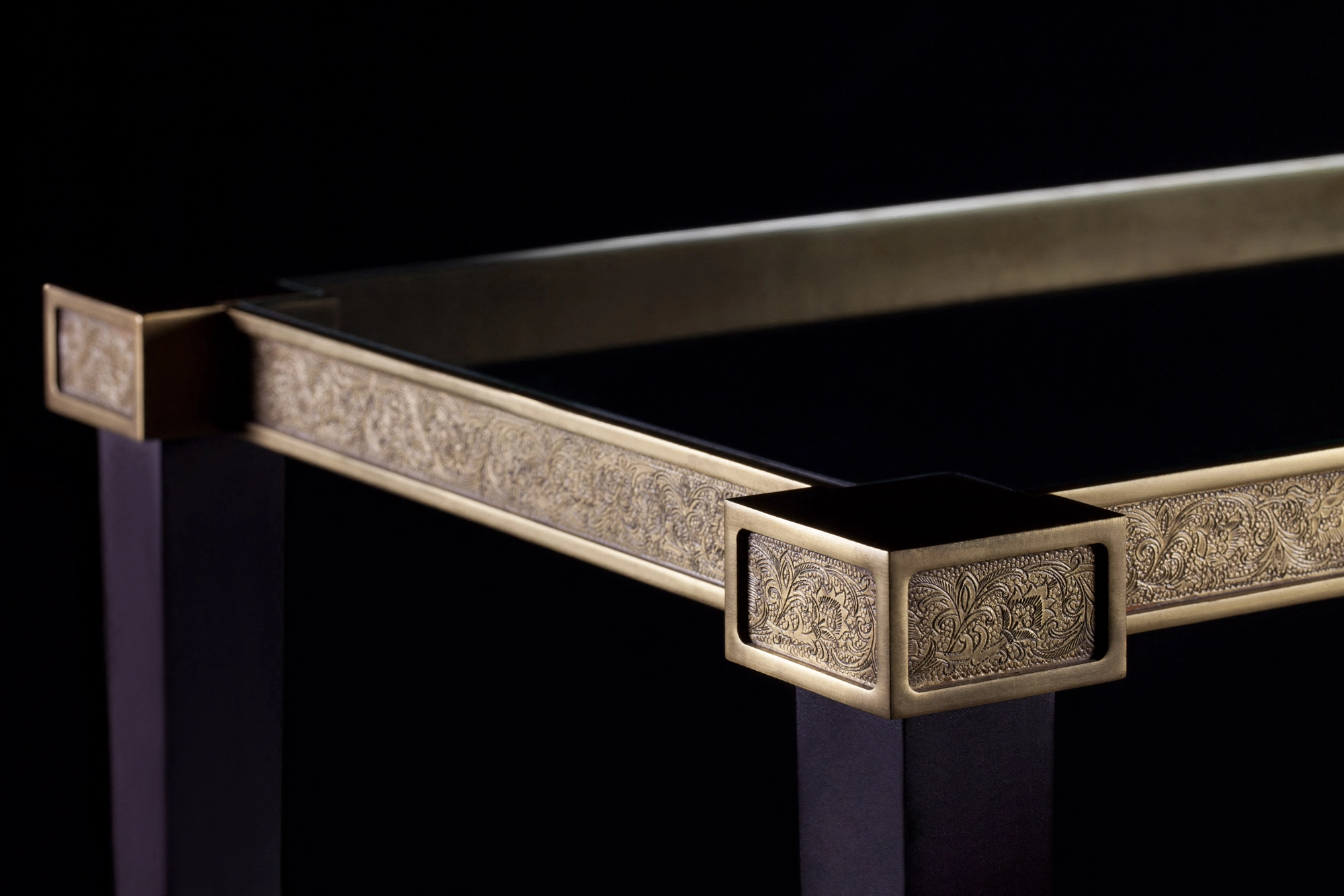 Corner detail of a glass console table with an engraved brass bracket, against a black background.